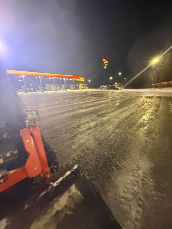 View from the cab of a snowplow clearing a snowy, icy gas station lot at night under bright overhead lights.