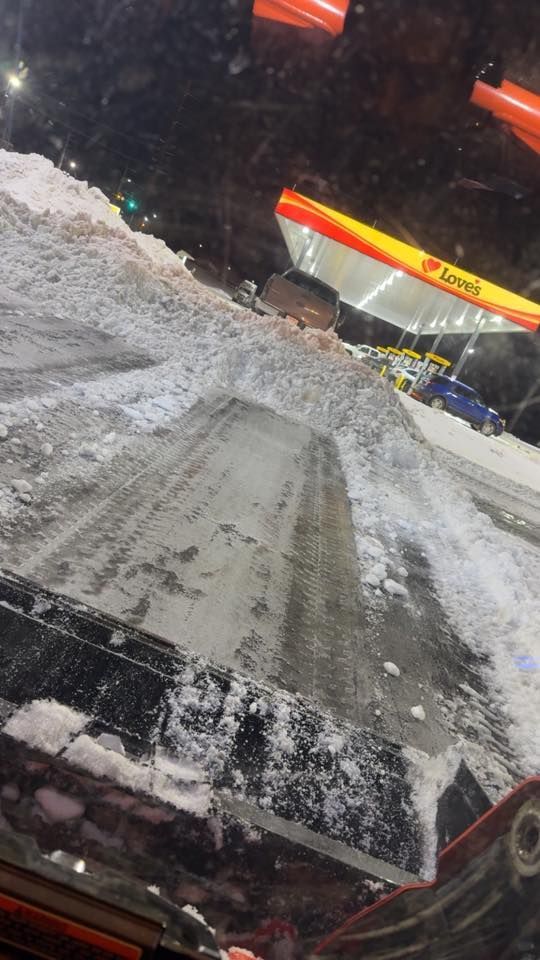 View from inside a vehicle at night, looking out at a snow-covered parking lot with a brightly lit Shell gas station.