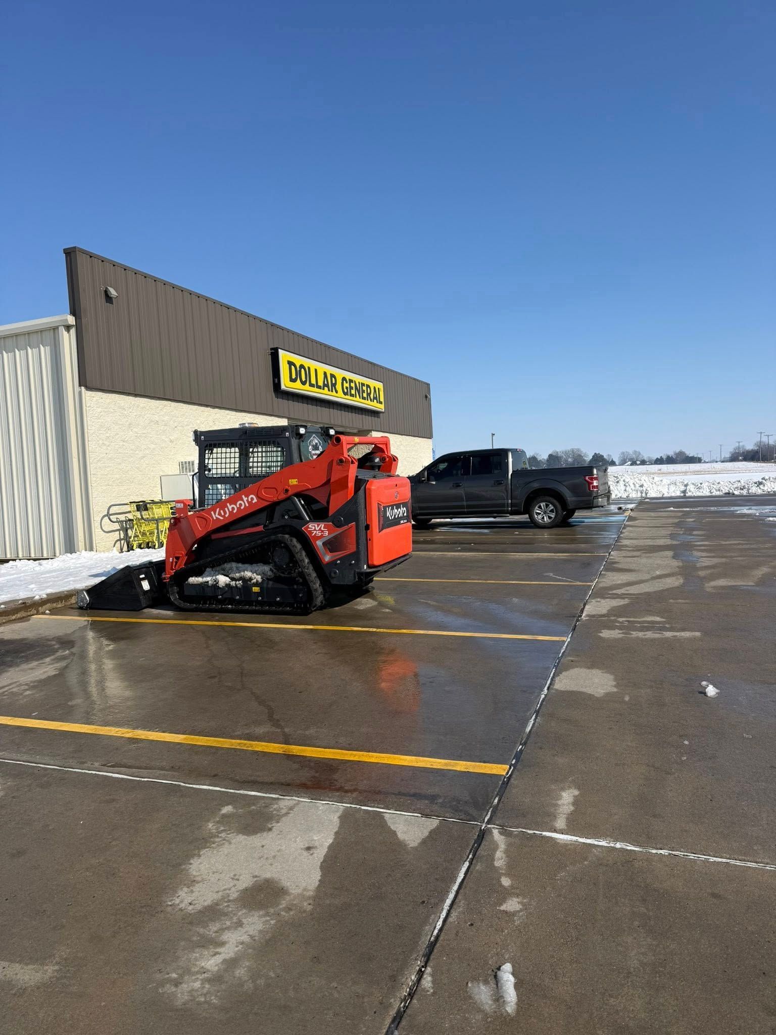 A red tracked skid steer parked in a marked lot in front of a building with a yellow sign under a clear blue sky.