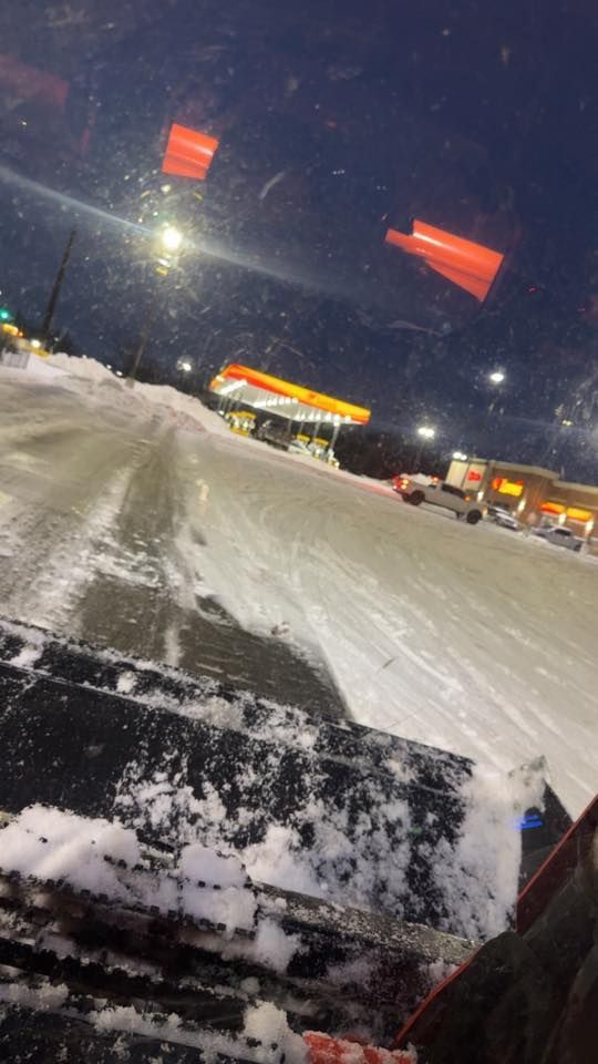 View from inside a vehicle at night, looking at a snow-covered parking lot with a gas station in the distance.