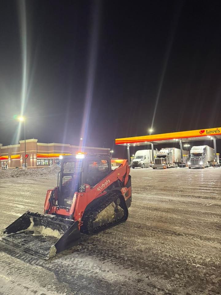 An orange Kubota skid-steer loader sits on a snowy lot at night in front of a brightly lit gas station and semi-trucks.