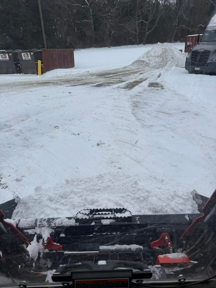 View from inside a machine, pushing a large blade through snow in a lot with dumpsters and a truck nearby.