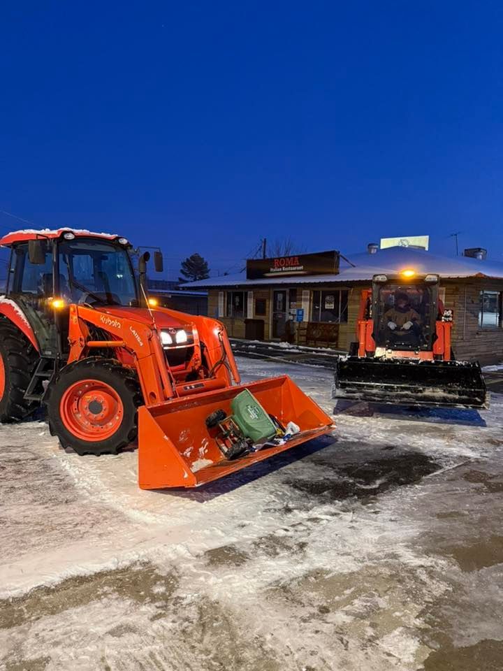 Two orange Kubota construction vehicles parked in a snow-covered parking lot in front of a building at dusk.
