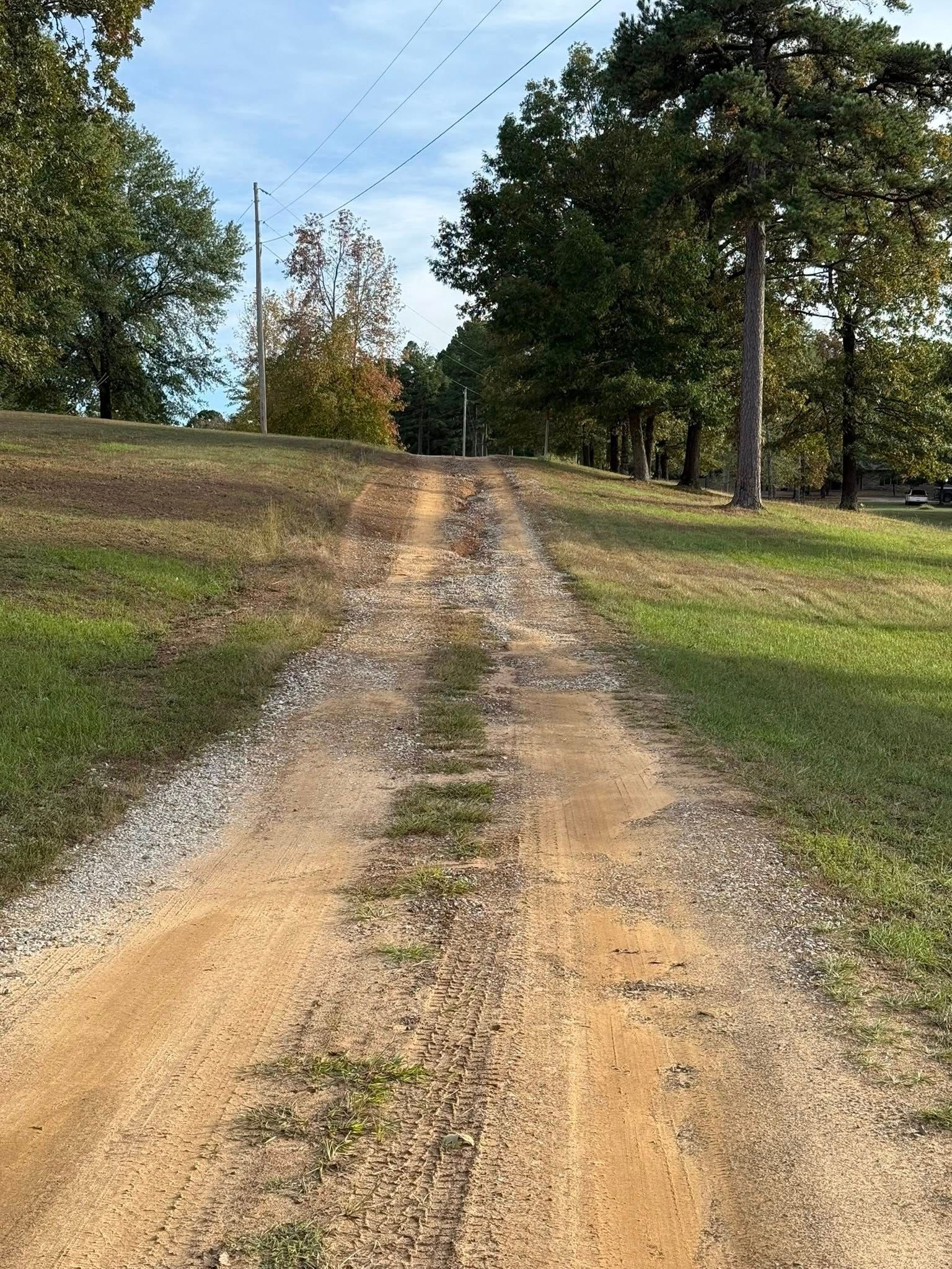 A gravel driveway leads uphill through a grassy landscape lined with trees under a blue sky.