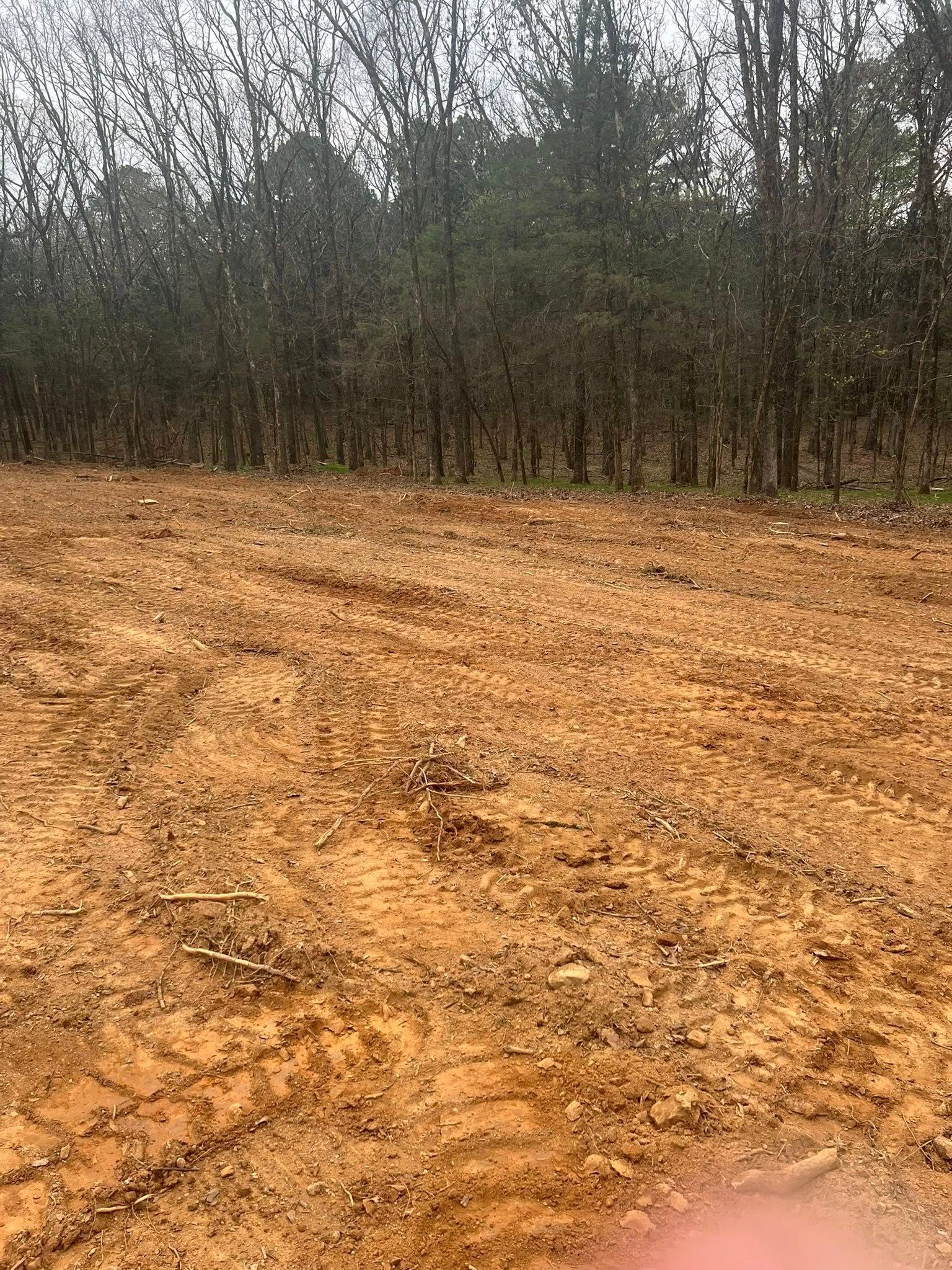A field of cleared, reddish-brown soil with tire tracks in the foreground, bordered by a dense line of trees in the back.