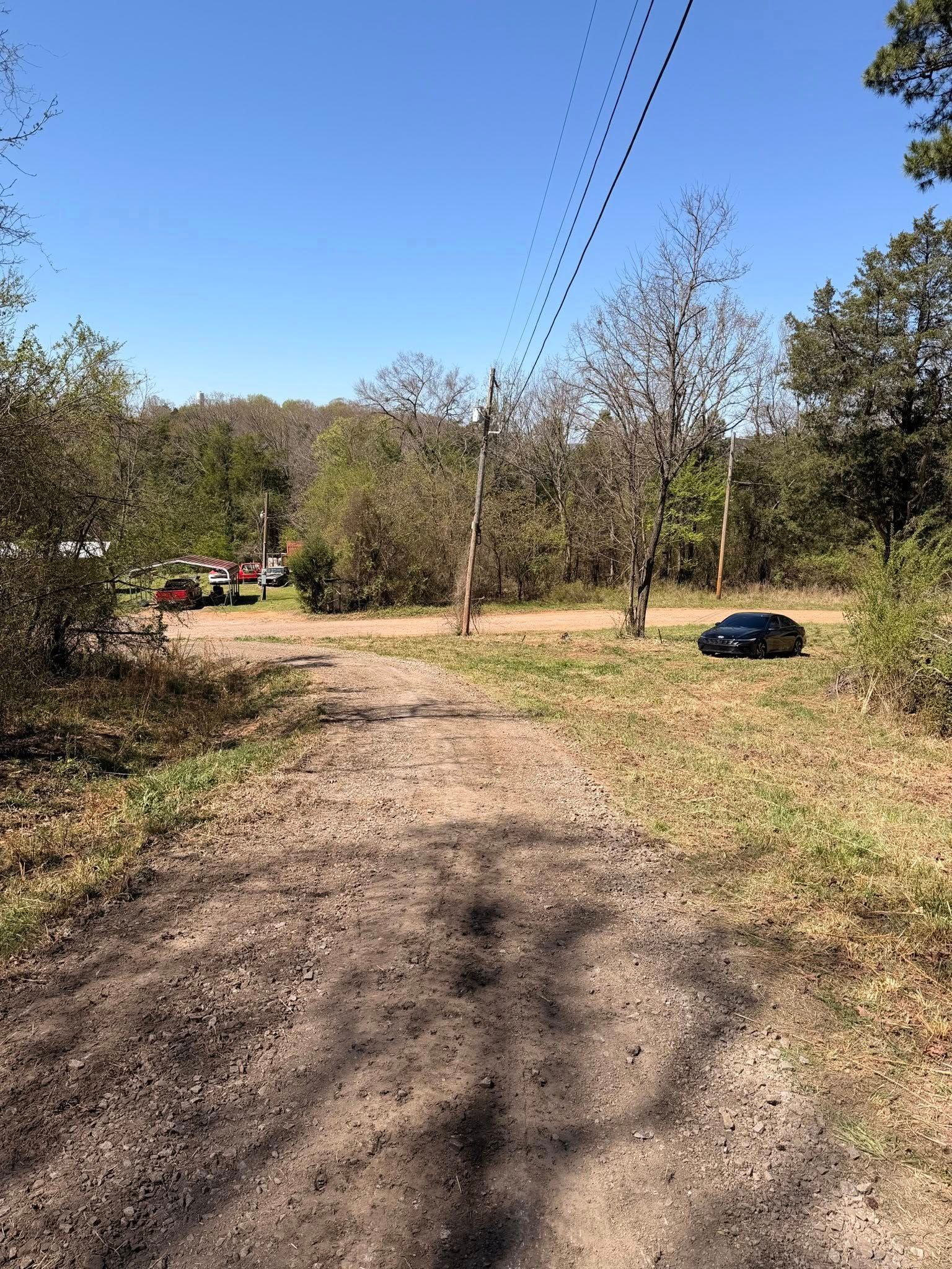 A dirt path leads to a grassy clearing with a dark car parked near trees under a bright blue sky.