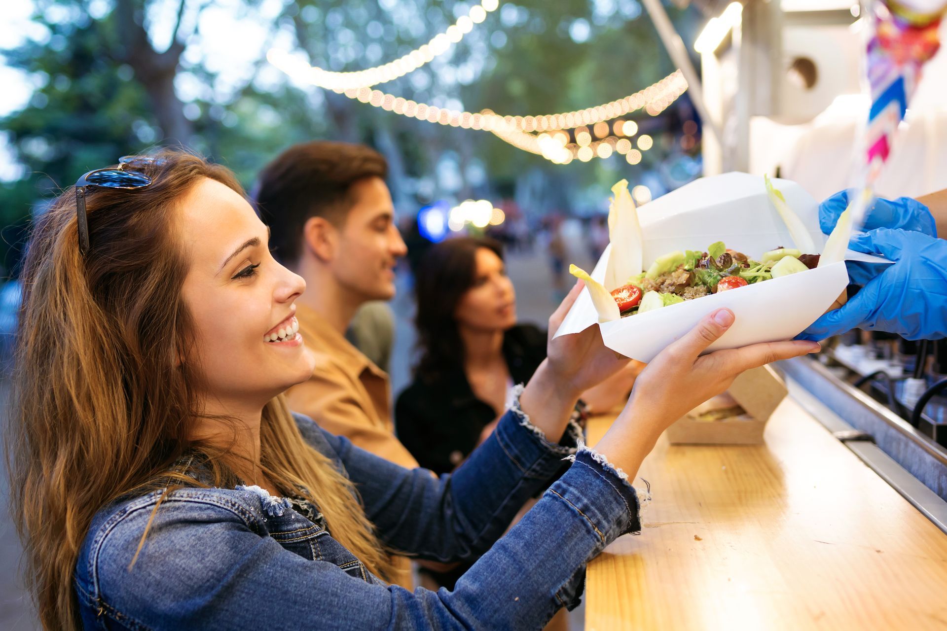 Woman smiling, receiving food from a food truck worker. Two other people are waiting in line behind her outdoors.