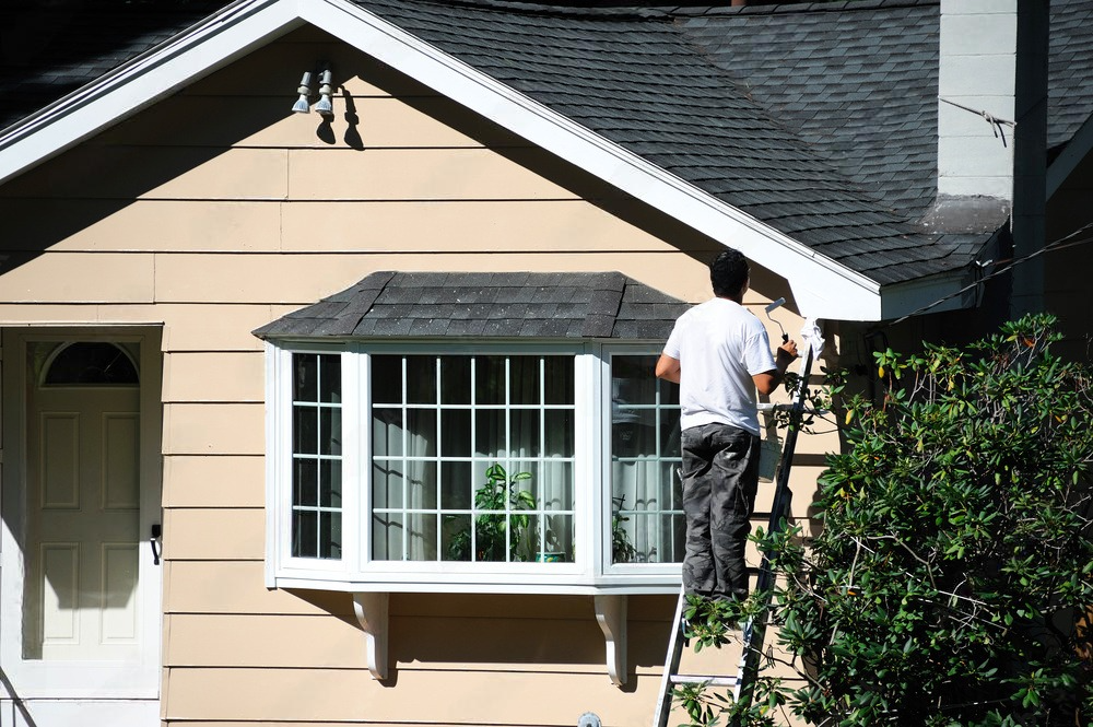 A man is standing on a ladder painting the side of a house.