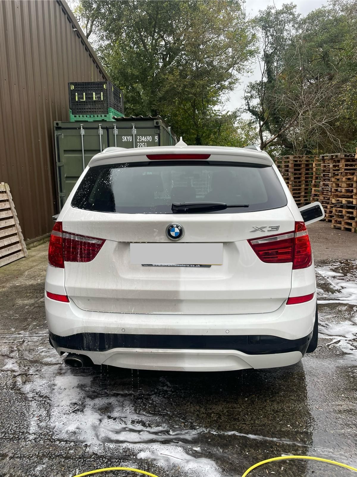 White BMW X3 SUV, rear view. Outdoors, wet surface, near a container and pallets.
