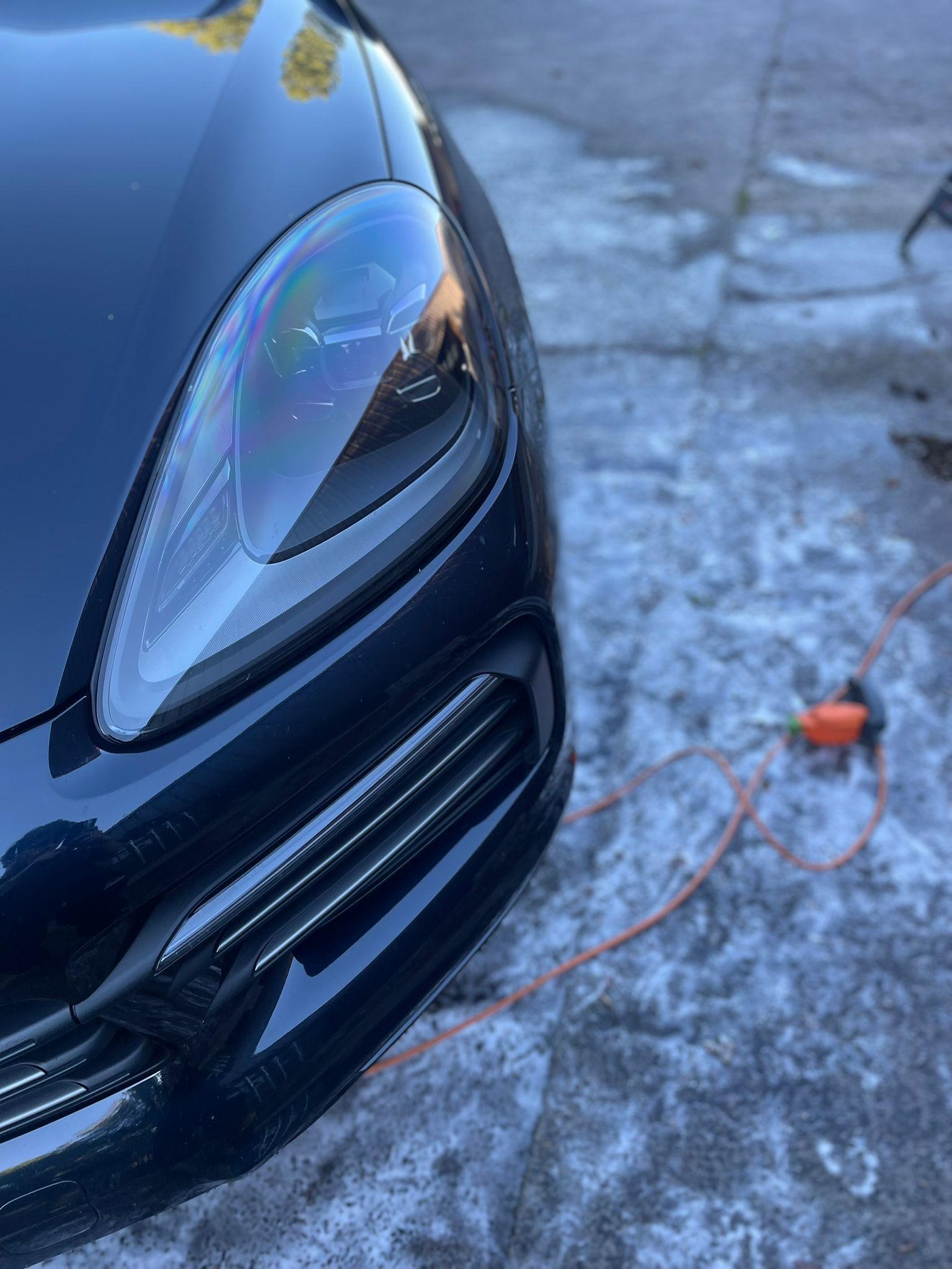 Black car headlight and grill, reflecting the surrounding environment, with an orange electrical tool and cord nearby.