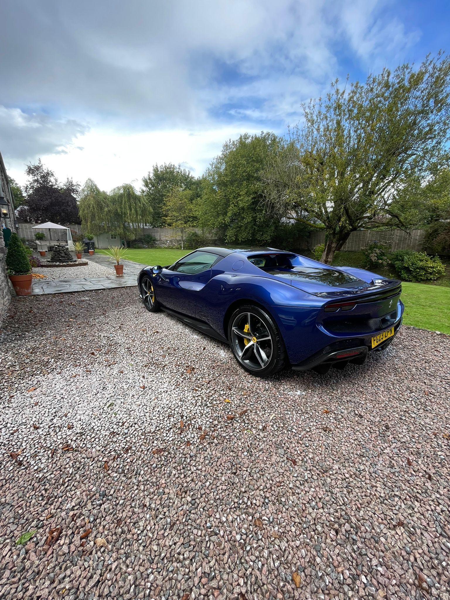 Blue sports car parked on gravel driveway, lush green background, overcast sky.