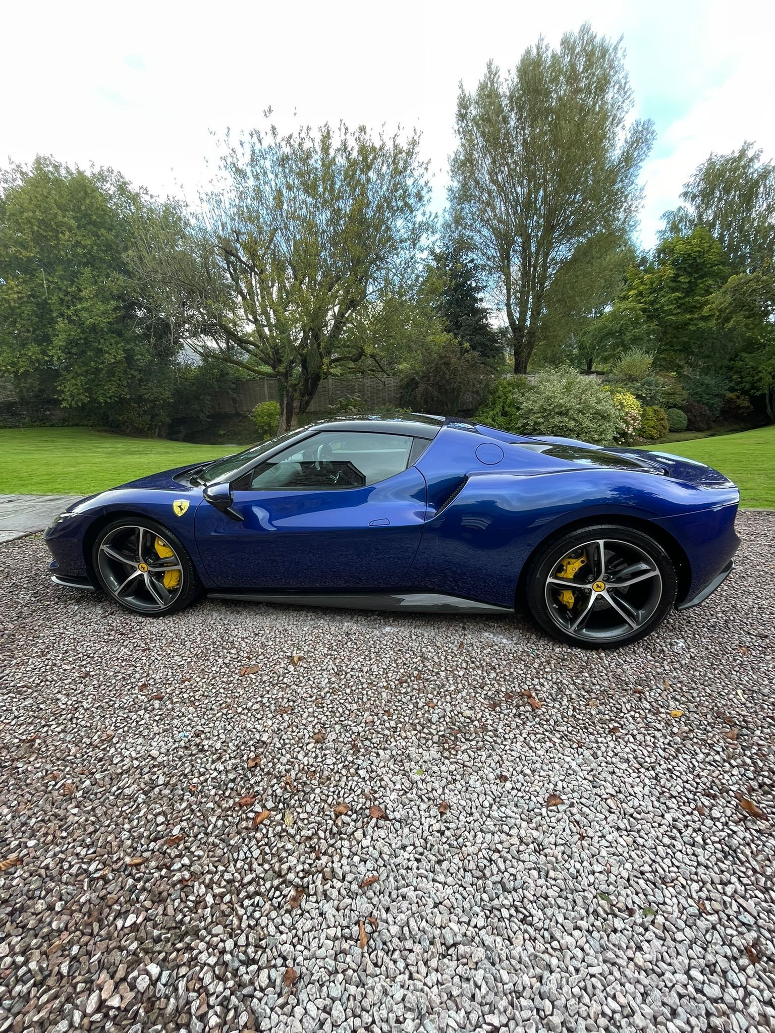 Blue Ferrari sports car parked on gravel, trees in the background, yellow brake calipers.