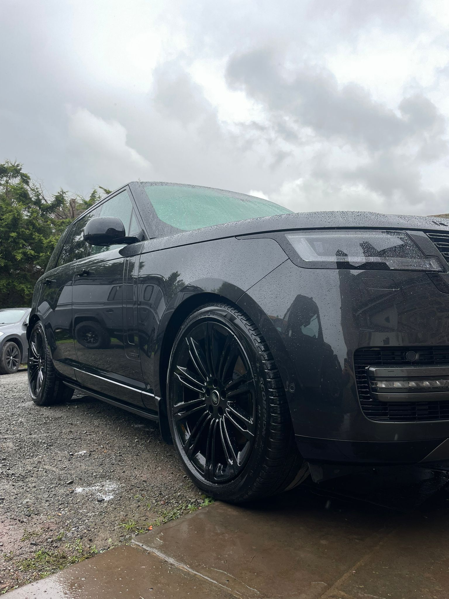 Dark gray Range Rover SUV parked, wet from rain, black wheels, under cloudy skies.