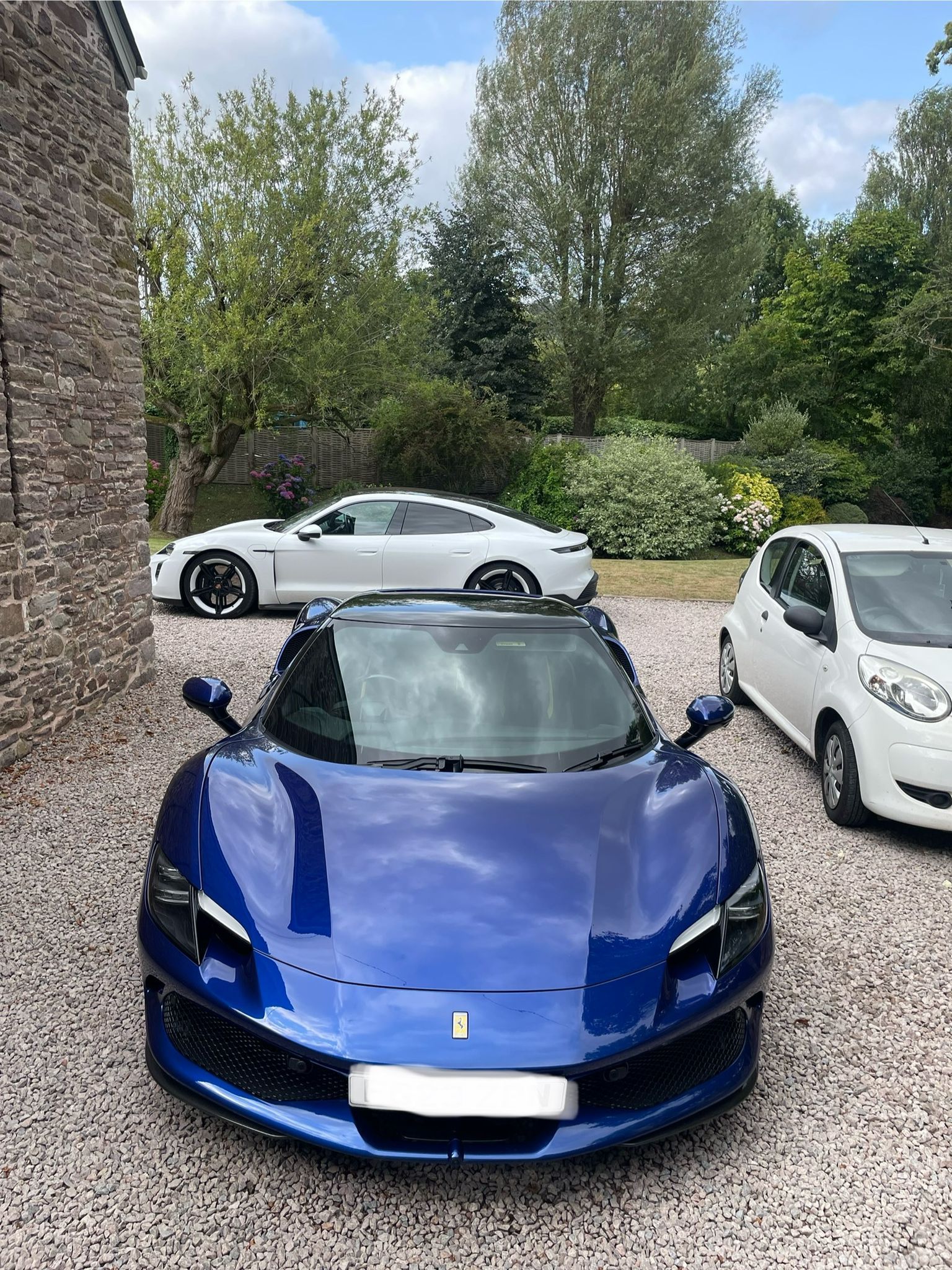 Blue Ferrari sports car parked on gravel, with white car and coupe in the background. Sunny day.
