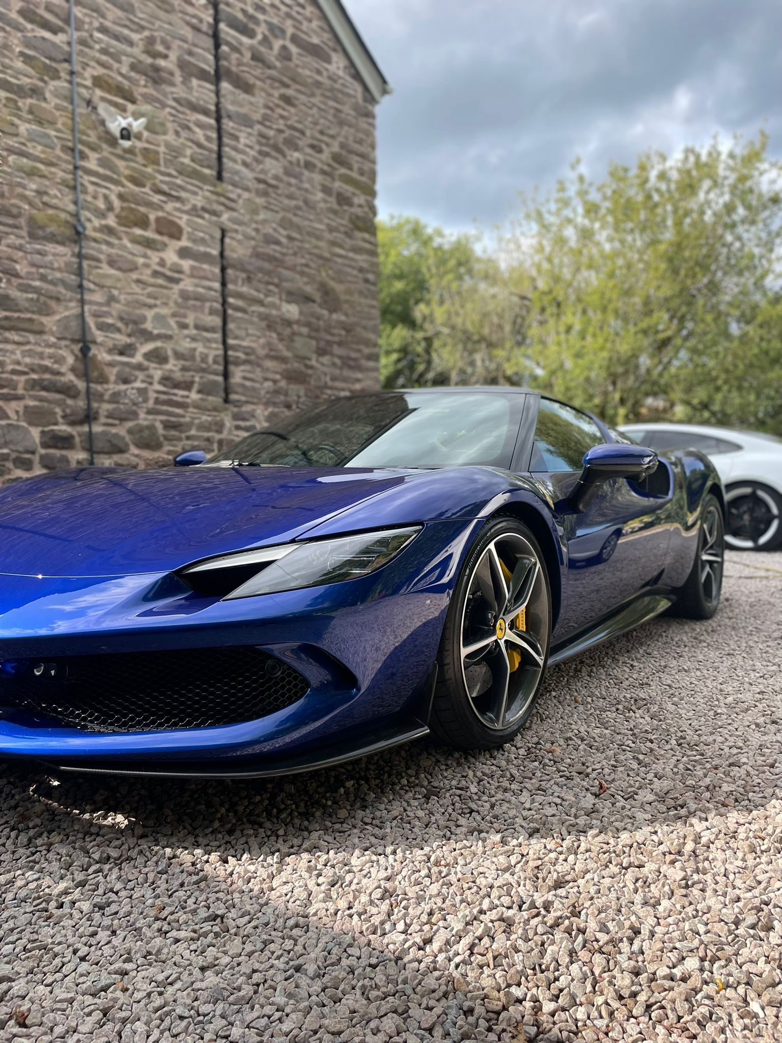 Blue sports car parked on gravel, near a stone building and a white car.