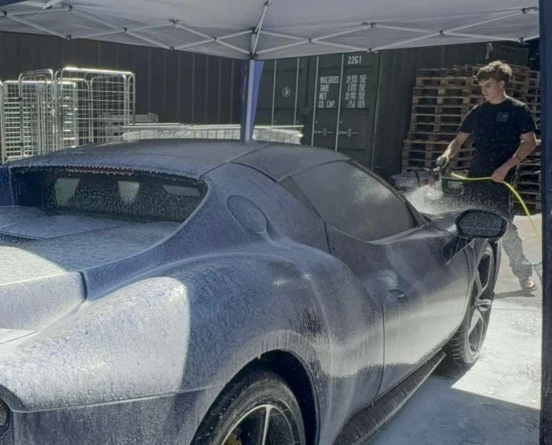 Person washing a blue sports car with foam. Outdoors, under a canopy.
