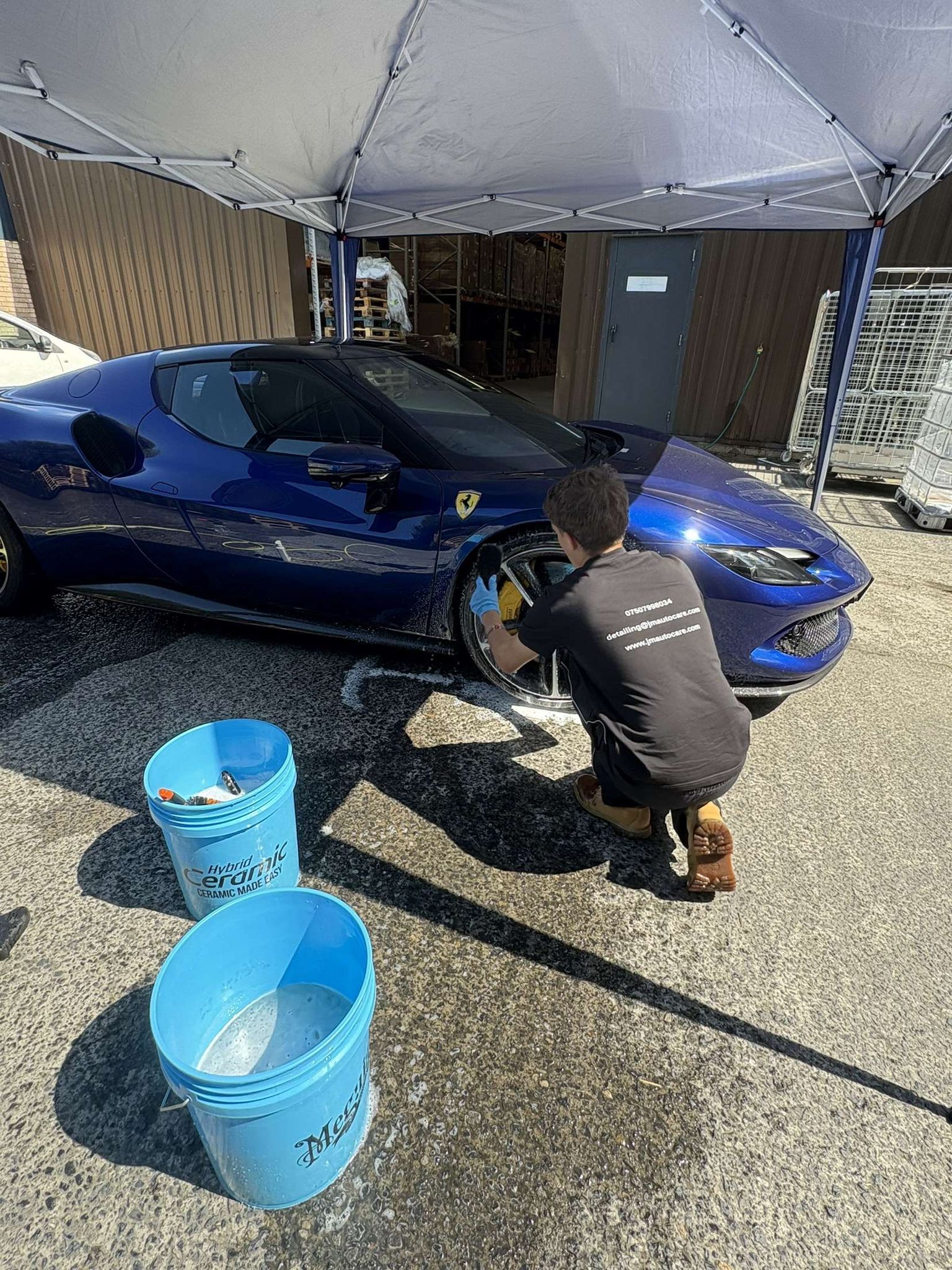 Person washing a blue sports car's wheel under a white canopy; two blue buckets nearby.