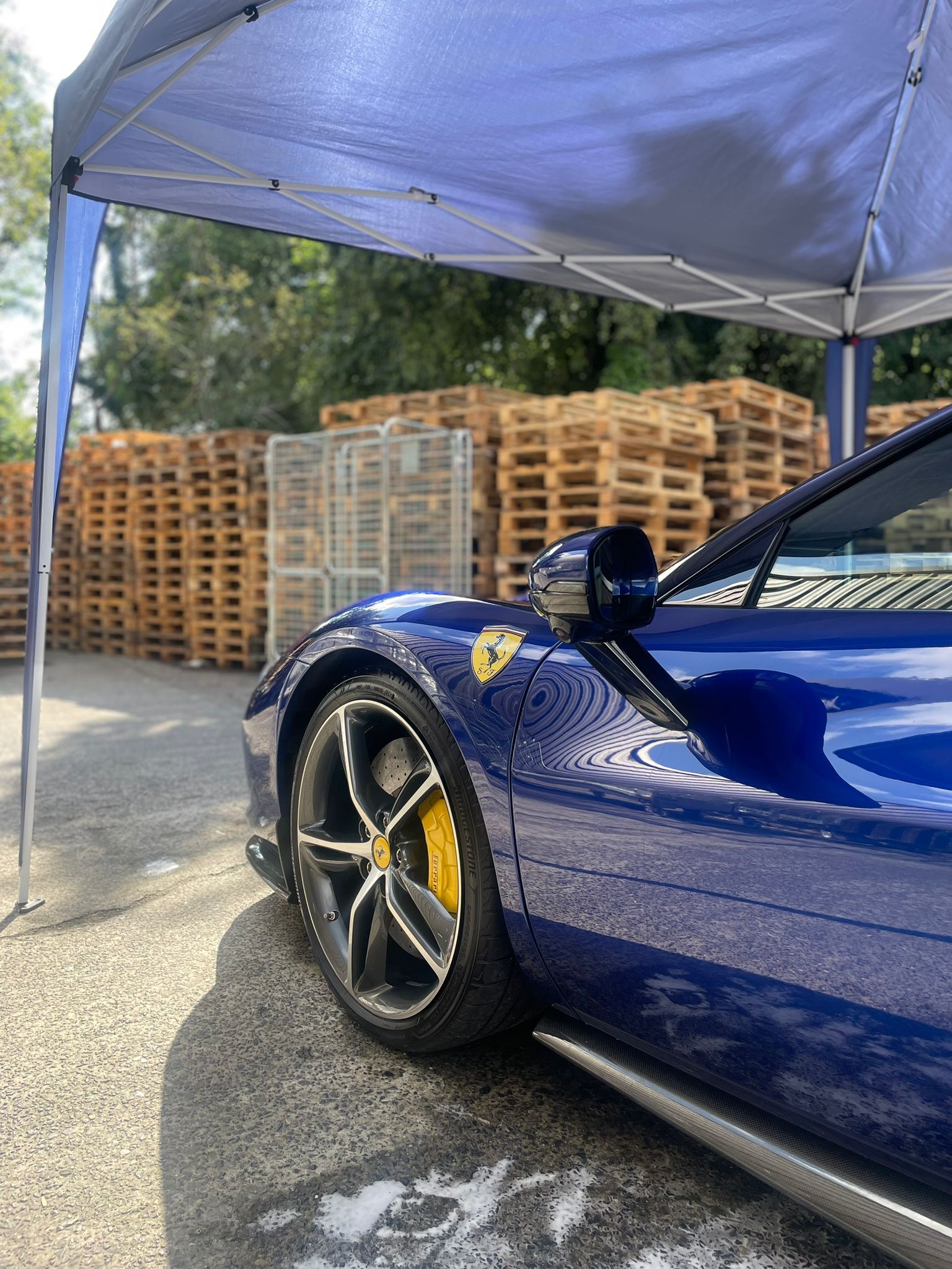 Blue Ferrari sports car parked under a canopy. Yellow brake calipers, pallets in the background.