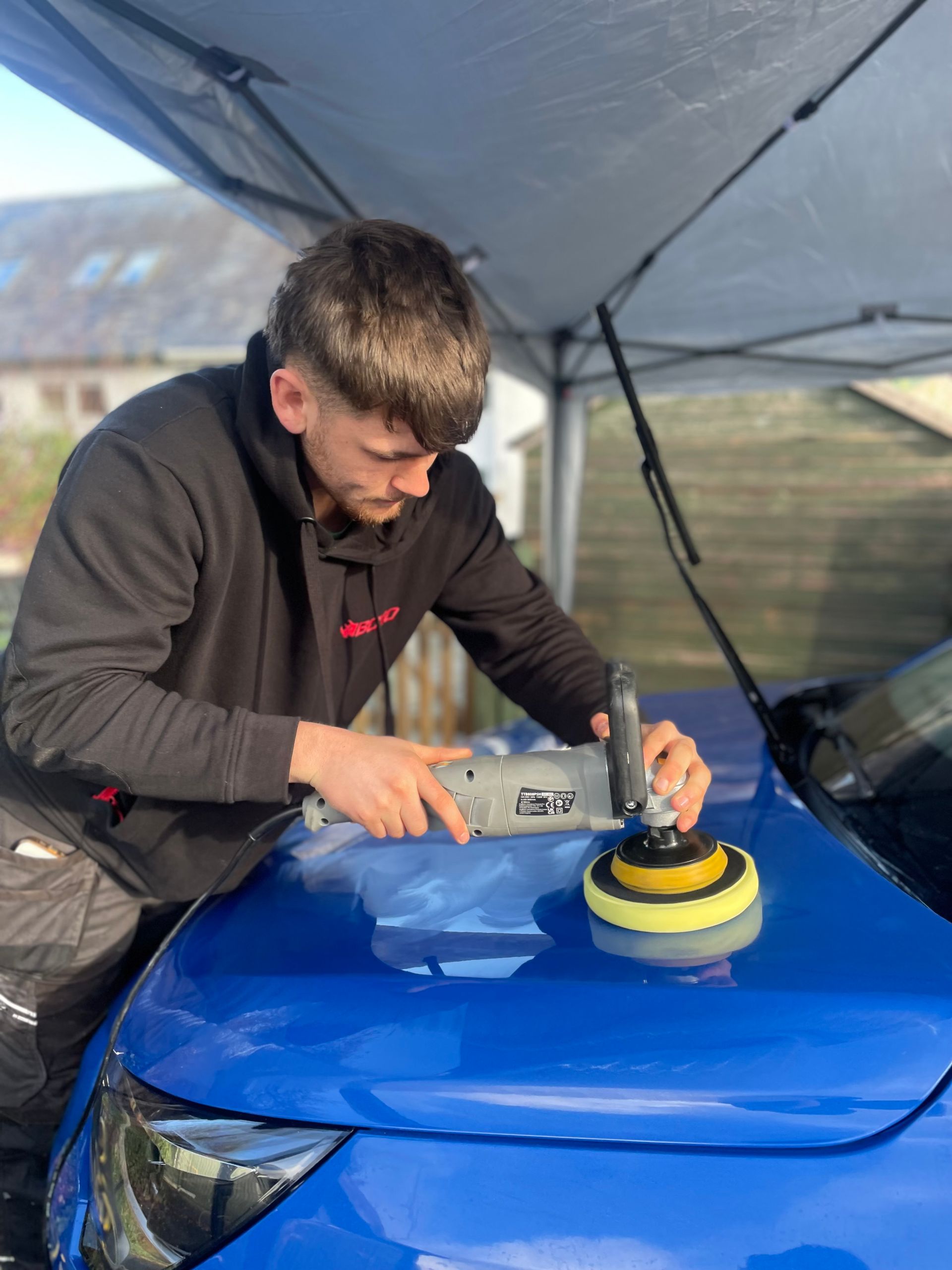 Man polishing a blue car with a buffer outside under a canopy.