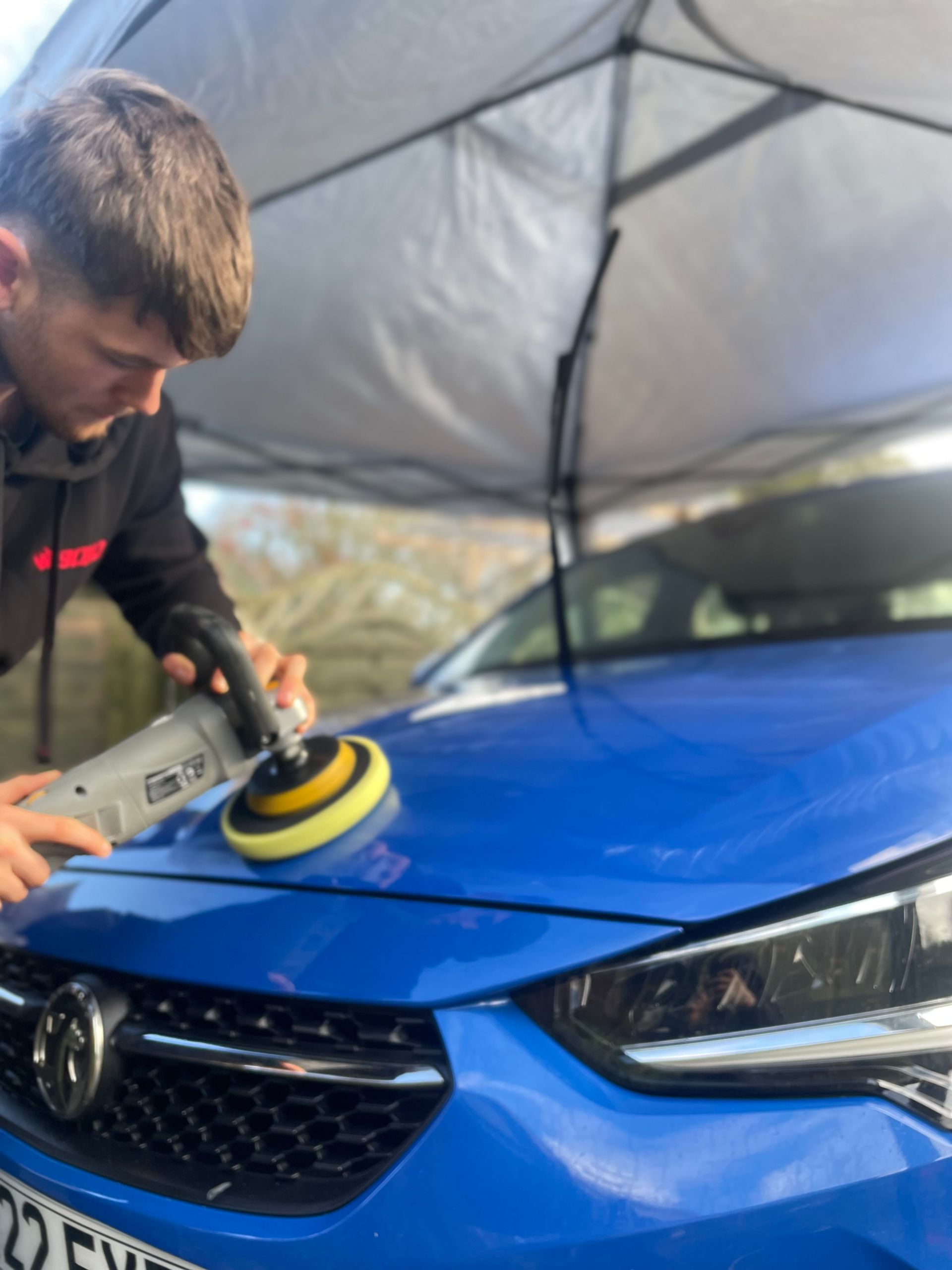 detailer polishing a blue car under a canopy. He is using a machine polisher to remove scratches.