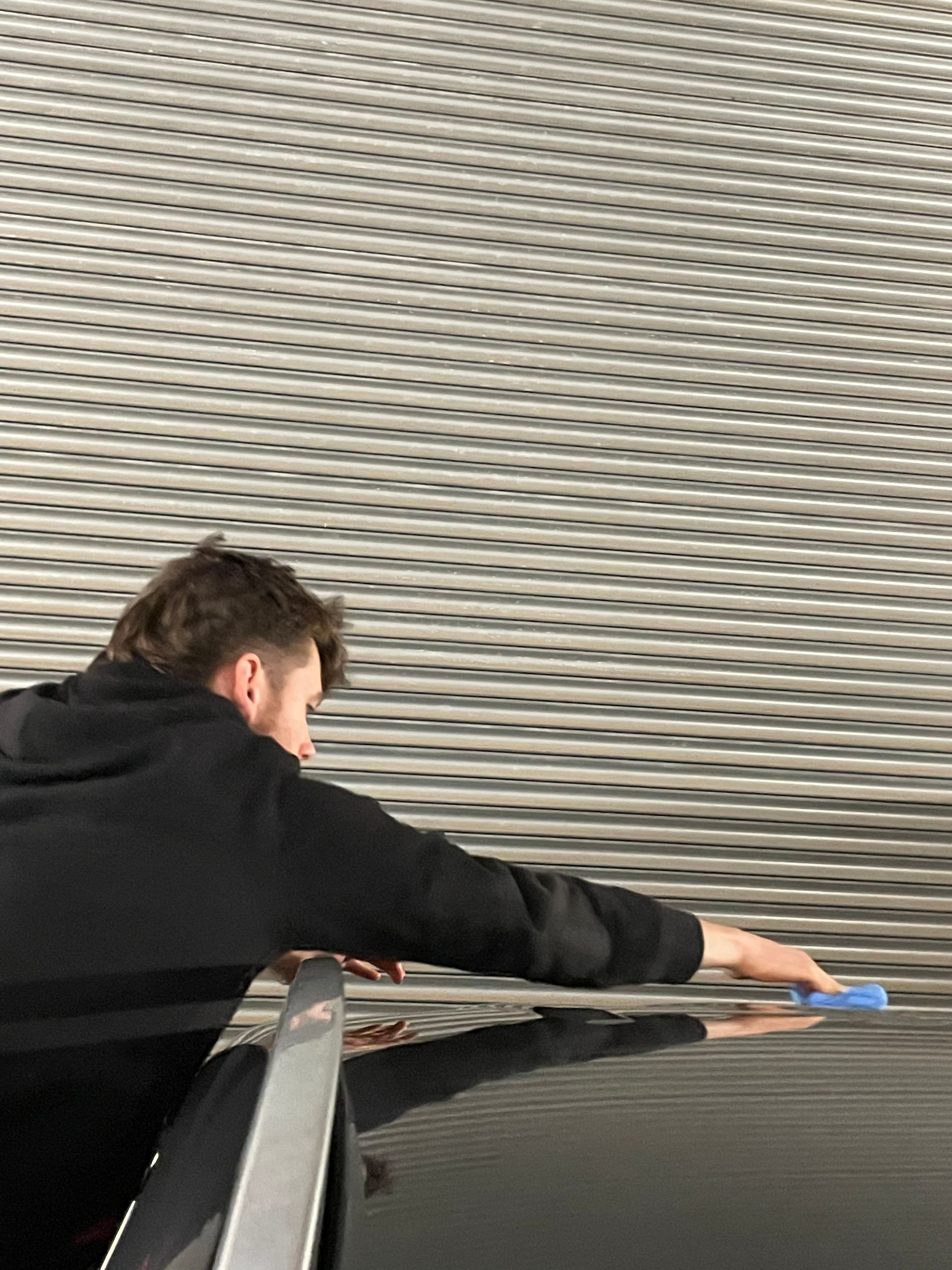 professional detailer in black hoodie using a blue applicator pad to apply ceramic coating to a grey car