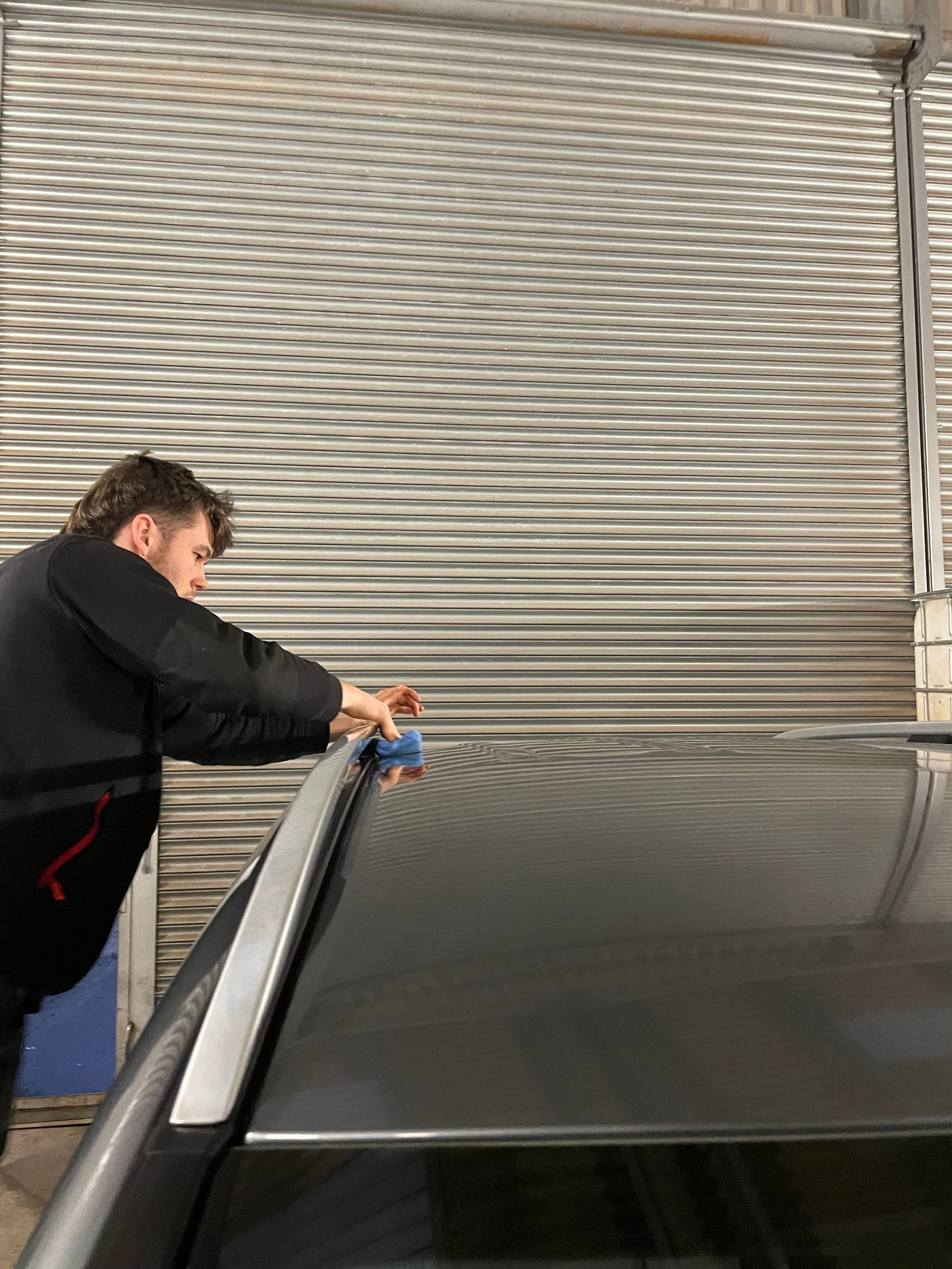 Professional detailer applying ceramic coating to a dark-colored car. Background is a metal shutter door.