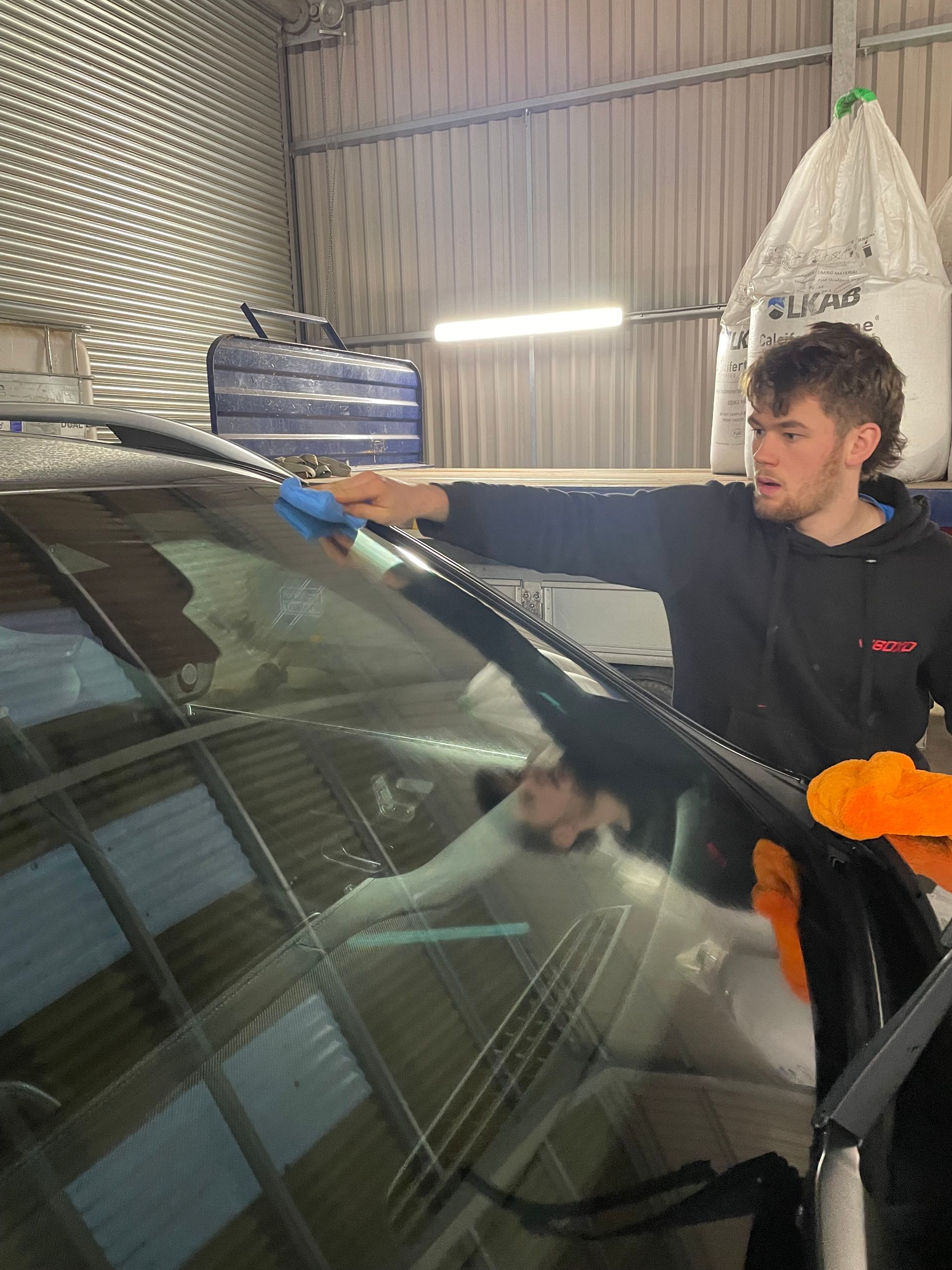 professional detailer using blue applicator pad to apply ceramic coating to windscreen of a grey luxury car