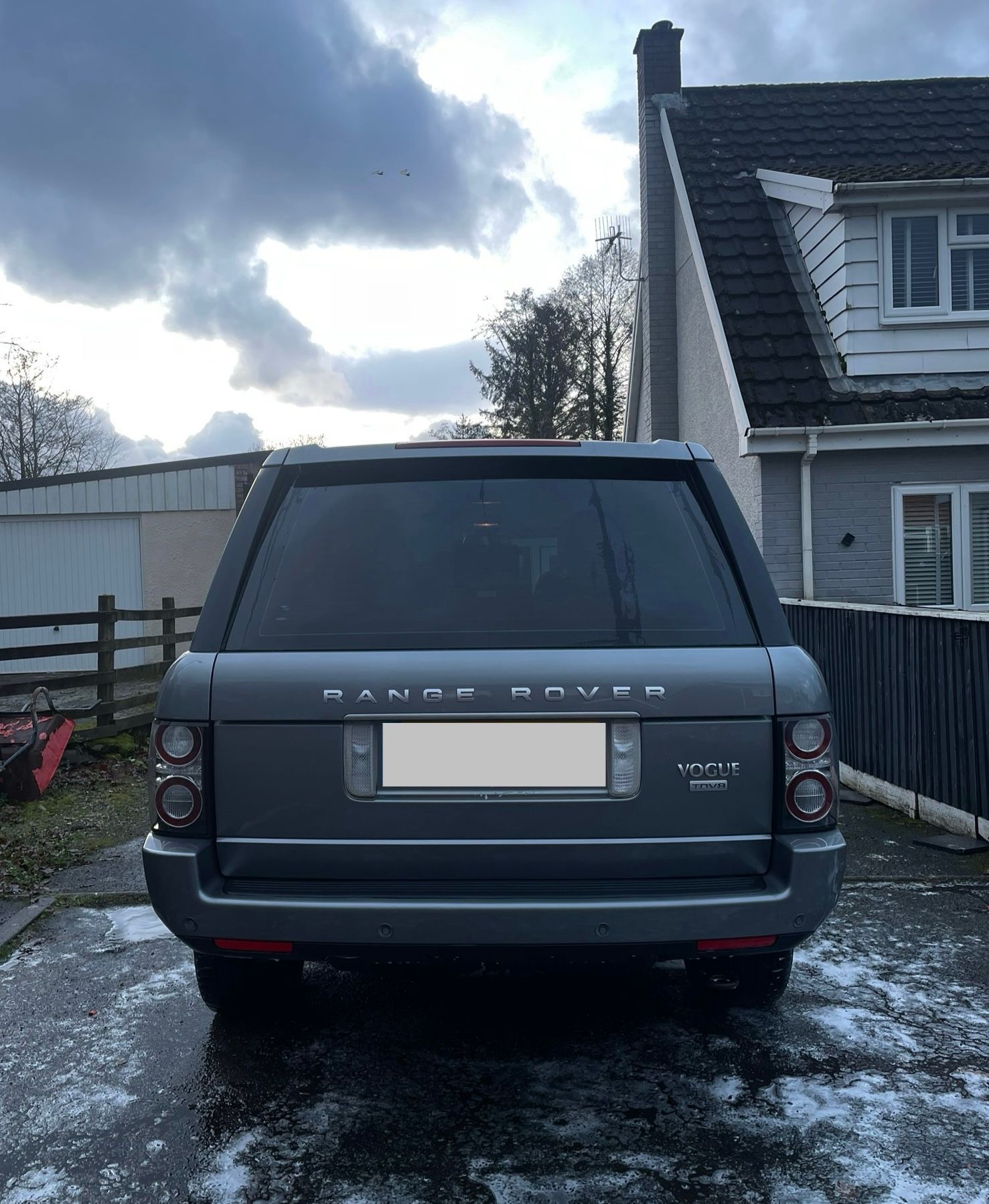 Gray Range Rover SUV parked on a wet surface, in front of a house, cloudy sky.