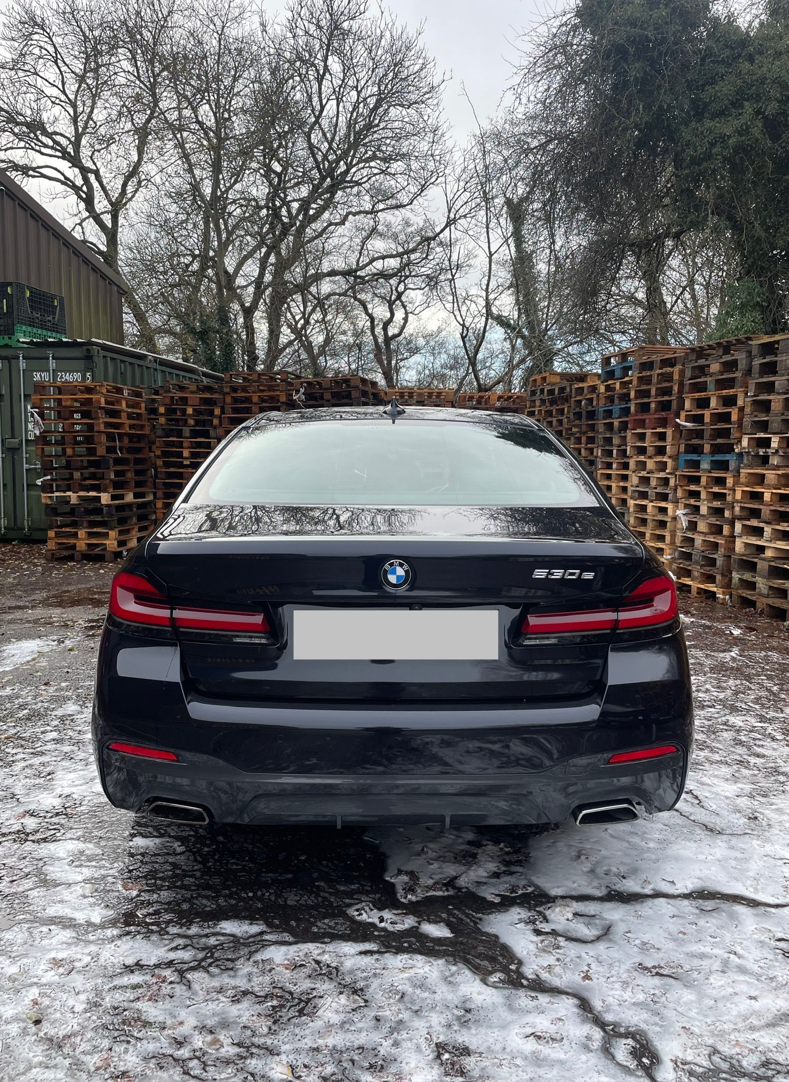 Black BMW sedan parked outdoors, rear view. Wet pavement, pallets and bare trees in the background.