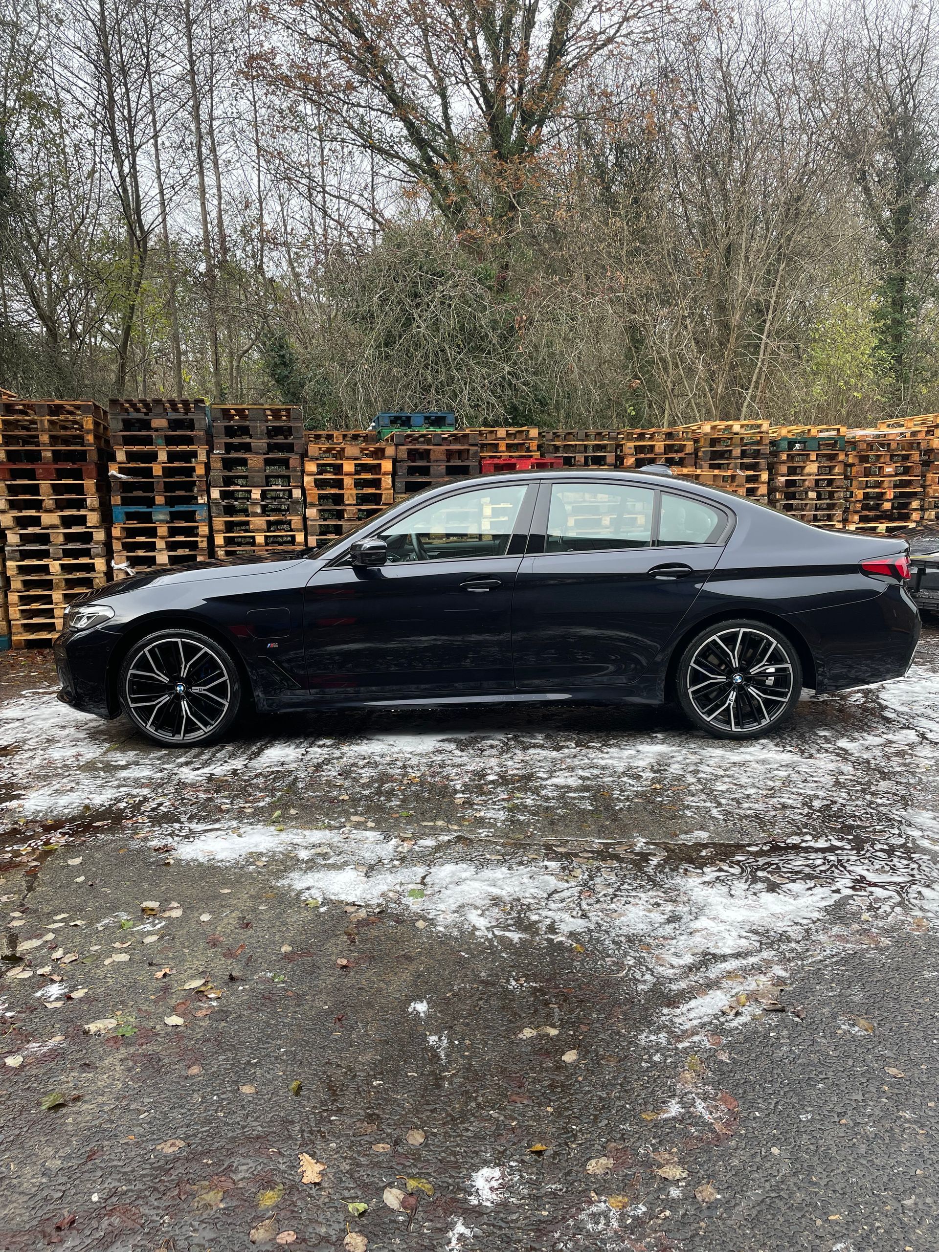 Black sedan parked on a snowy surface near wooden pallets and trees.