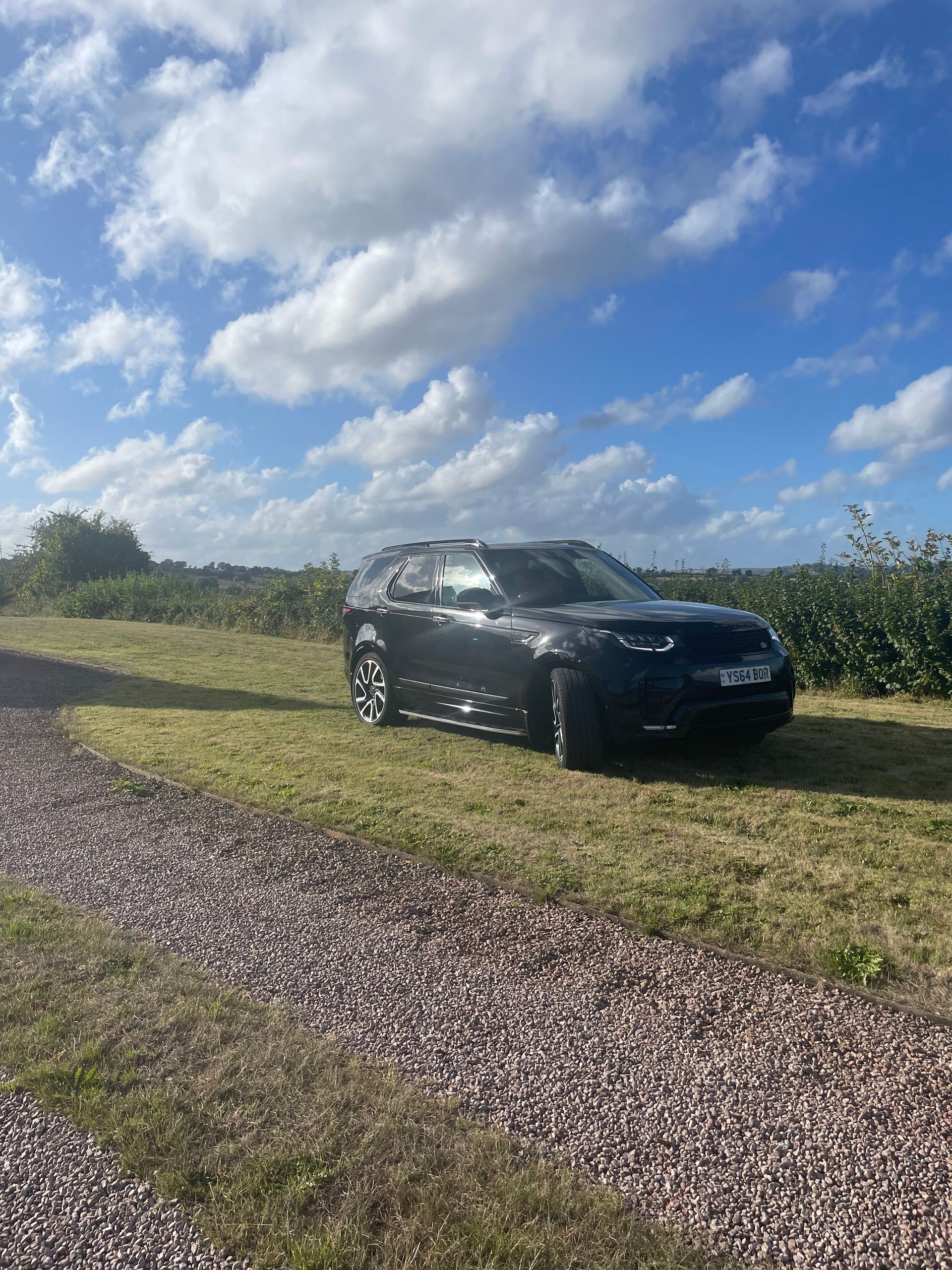 Black SUV parked on grass near a gravel path, under a cloudy blue sky.