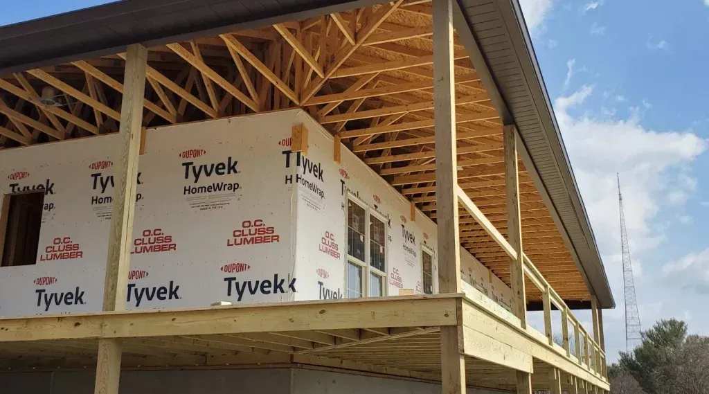 A low-angle view of a house under construction with white Tyvek house wrap, wooden deck framing, and exposed roof trusses.