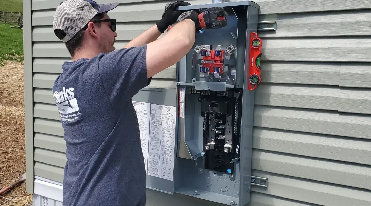 A person in a gray t-shirt and cap installs an electrical meter box on the side of a building with light green siding.
