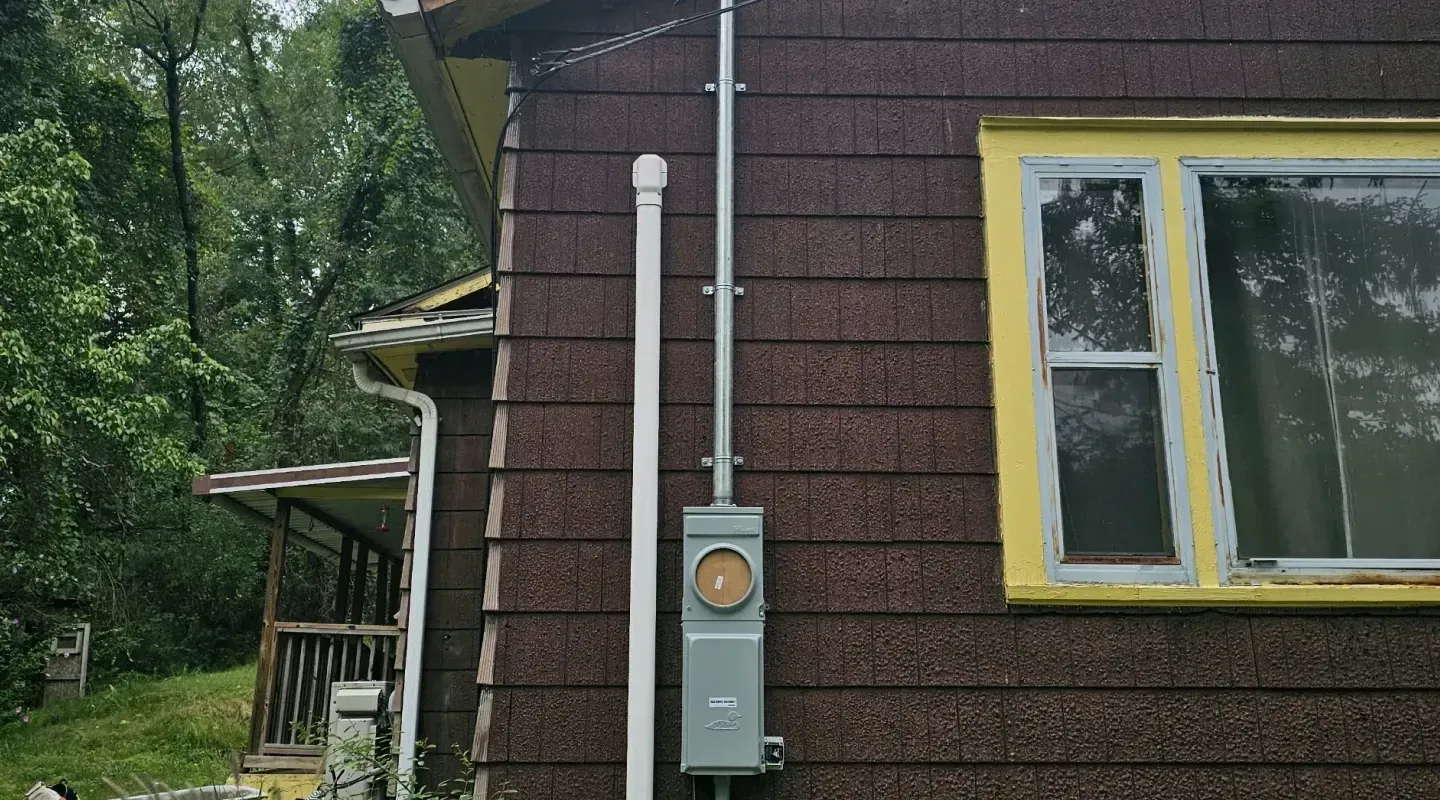 An exterior wall of a house with a gray electrical meter box mounted on it, with conduit running up to the roofline.