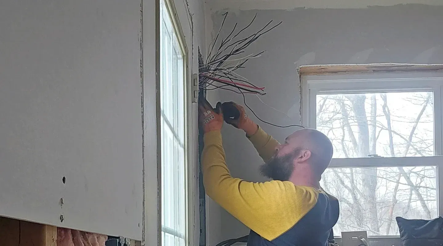 A person in a yellow and blue shirt uses a rotary tool to cut drywall near a bundle of electrical wires by a window.