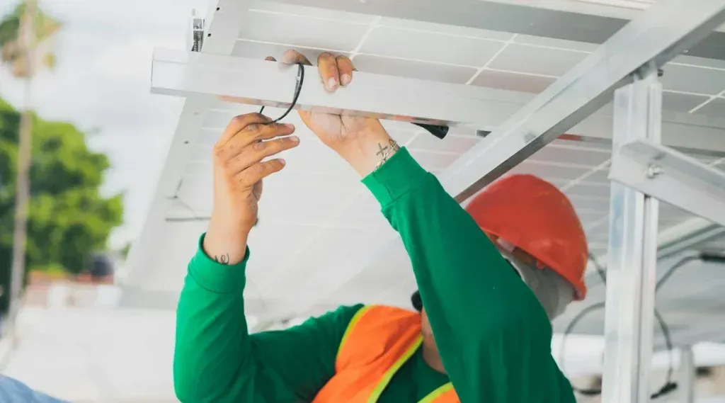 A worker in an orange hard hat and safety vest secures wiring to the metal frame of a solar panel installation.