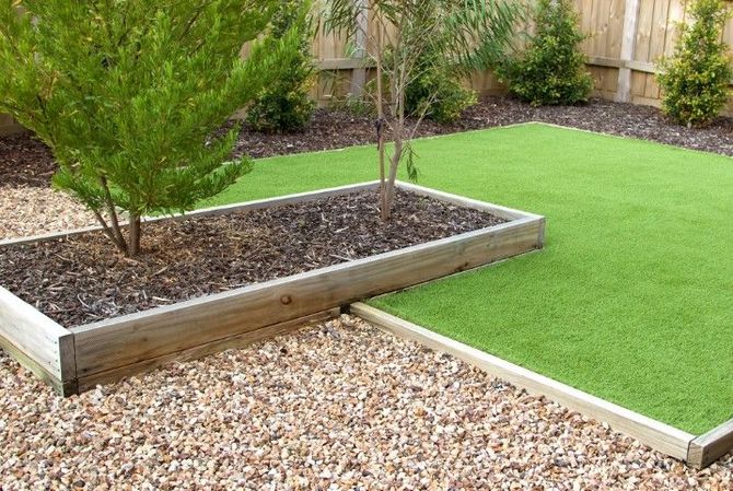 A garden featuring a rectangular wooden planter with mulch and trees, set beside a patch of green synthetic turf.