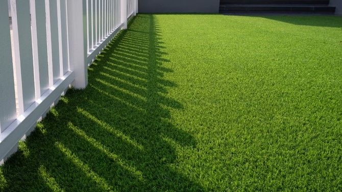 A white picket fence casts a long, geometric shadow across a vibrant green patch of artificial grass.