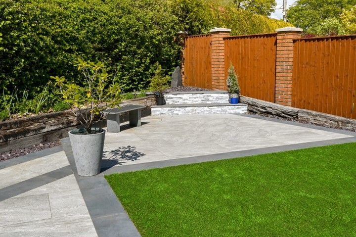 A modern backyard patio with grey pavers, a green lawn, a stone bench, and a raised planter bed against a wooden fence.