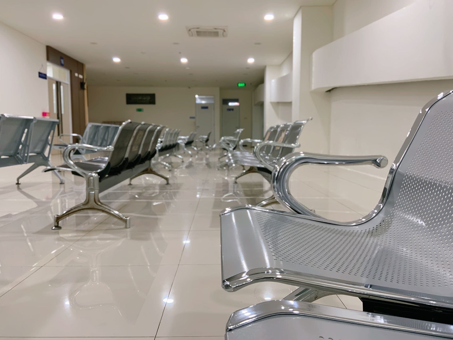 Rows of empty metallic waiting room chairs in a clean, modern, white-tiled hallway with overhead lighting.