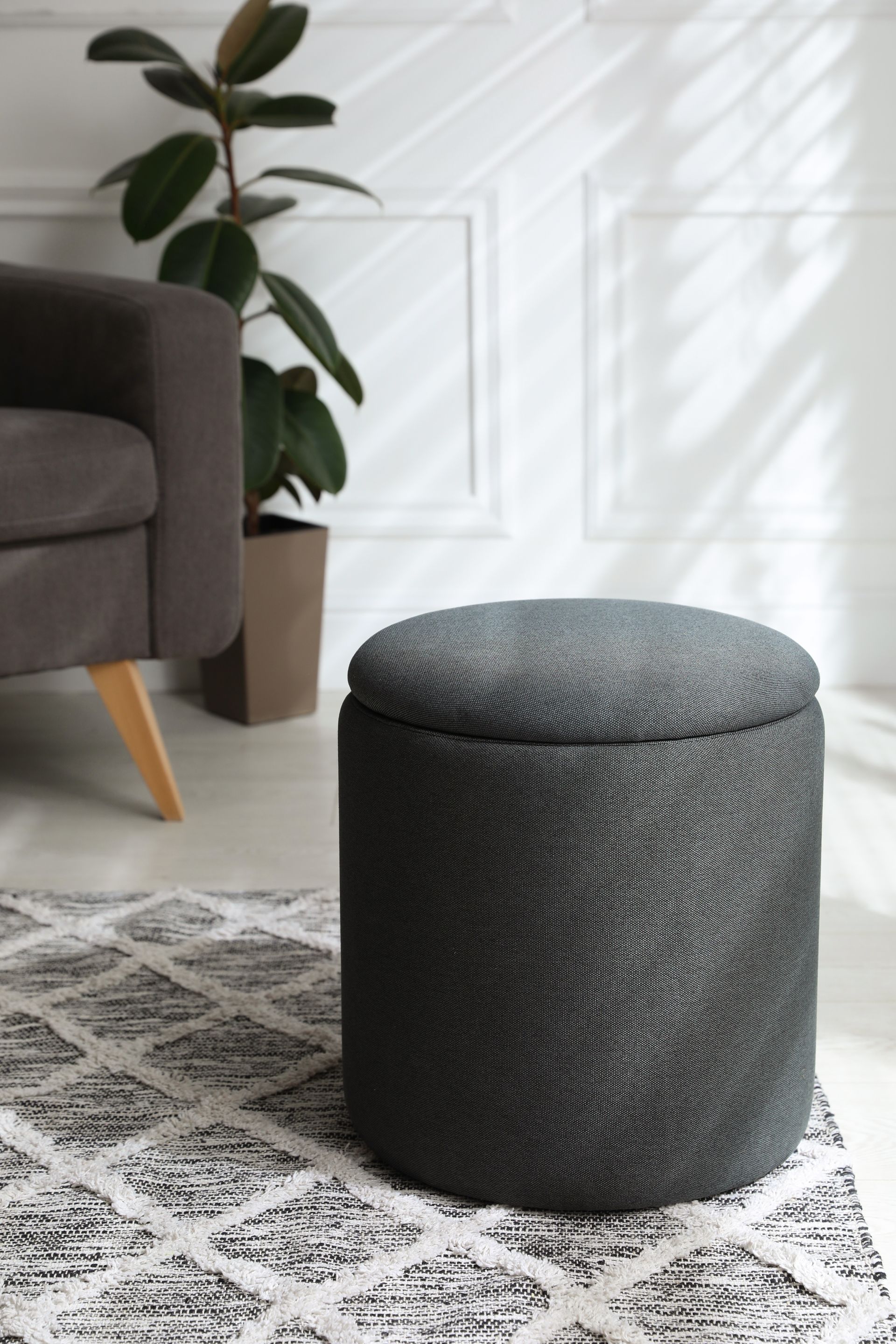 A textured dark gray cylindrical ottoman sits on a patterned rug next to a potted plant and a dark sofa.