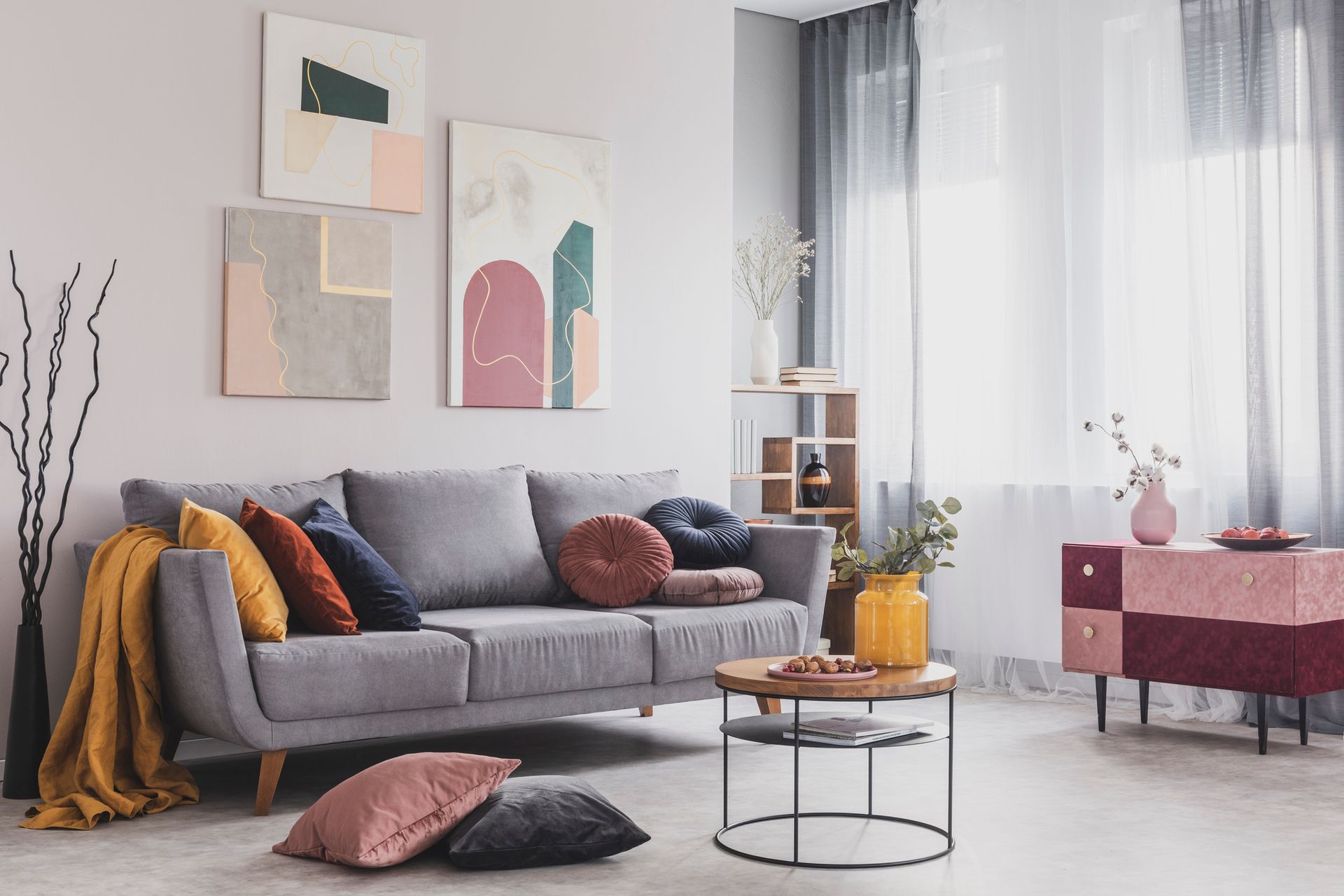 A modern living room with a grey sofa, colorful throw pillows, abstract art, a round wooden coffee table, and a cabinet.