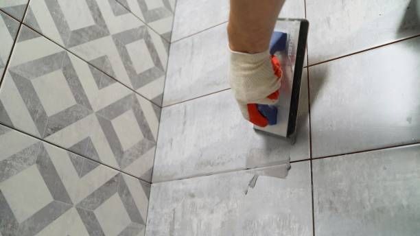 A hand in a glove uses a rubber grout float to spread grey grout over floor tiles.