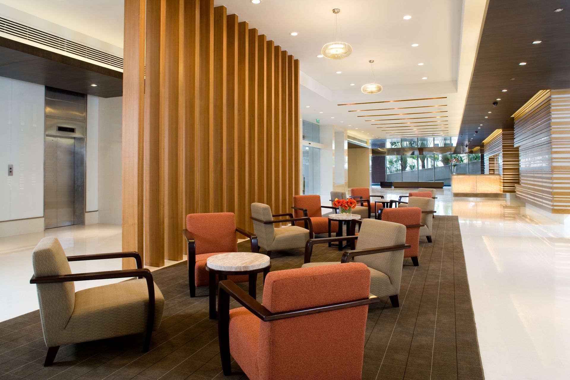 Modern hotel lobby with wood paneled walls, comfortable armchairs, and elevator access on a carpeted seating area.