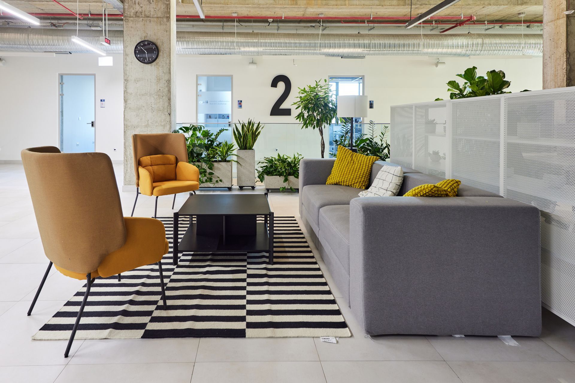A modern office lounge with a grey sofa, two mustard chairs, a coffee table on a striped rug, and indoor plants.