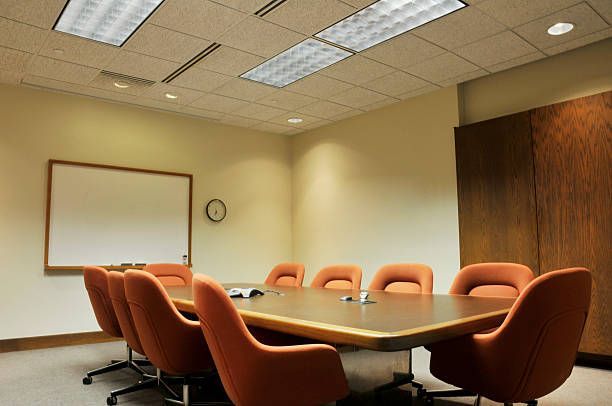 A conference room with a large wooden table, orange chairs, a blank whiteboard, and overhead recessed lighting.