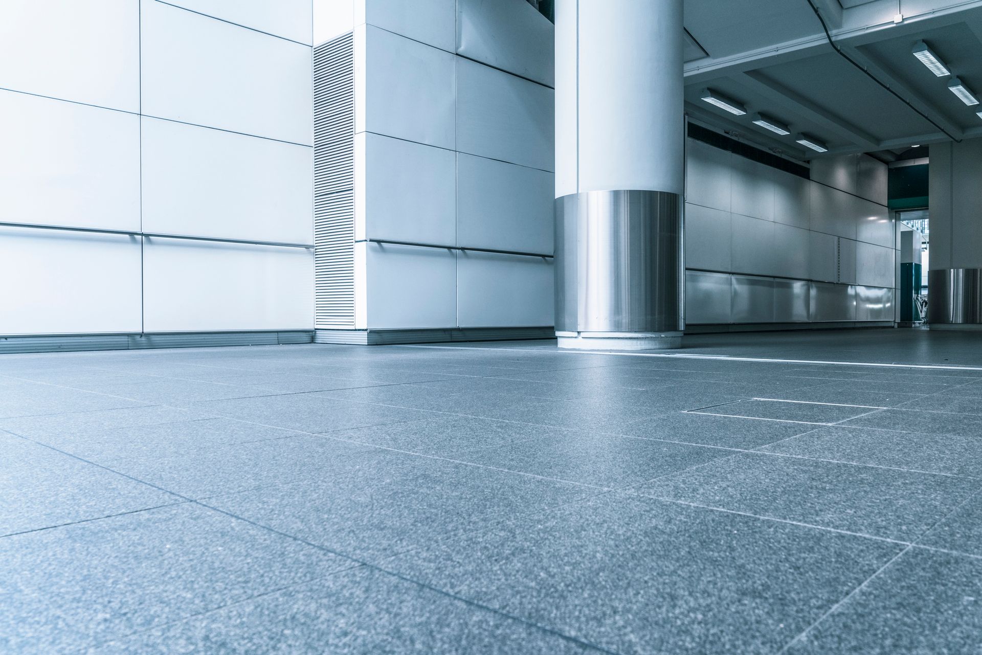 A modern, brightly lit indoor hallway with polished grey tiled floors, white paneled walls, and a large metallic pillar.