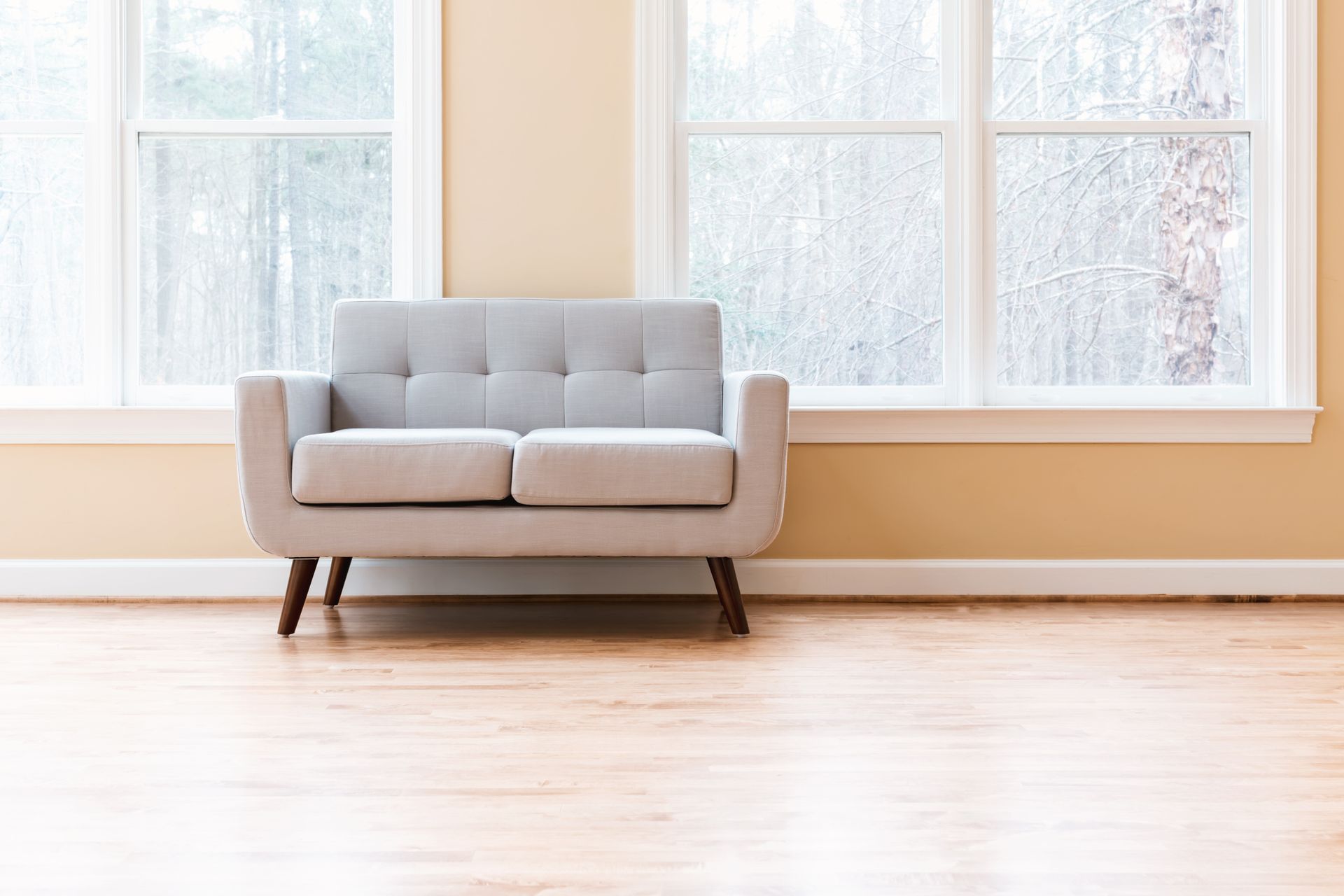 A light gray loveseat sits on a light wood floor in front of a tan wall with two large windows overlooking trees.