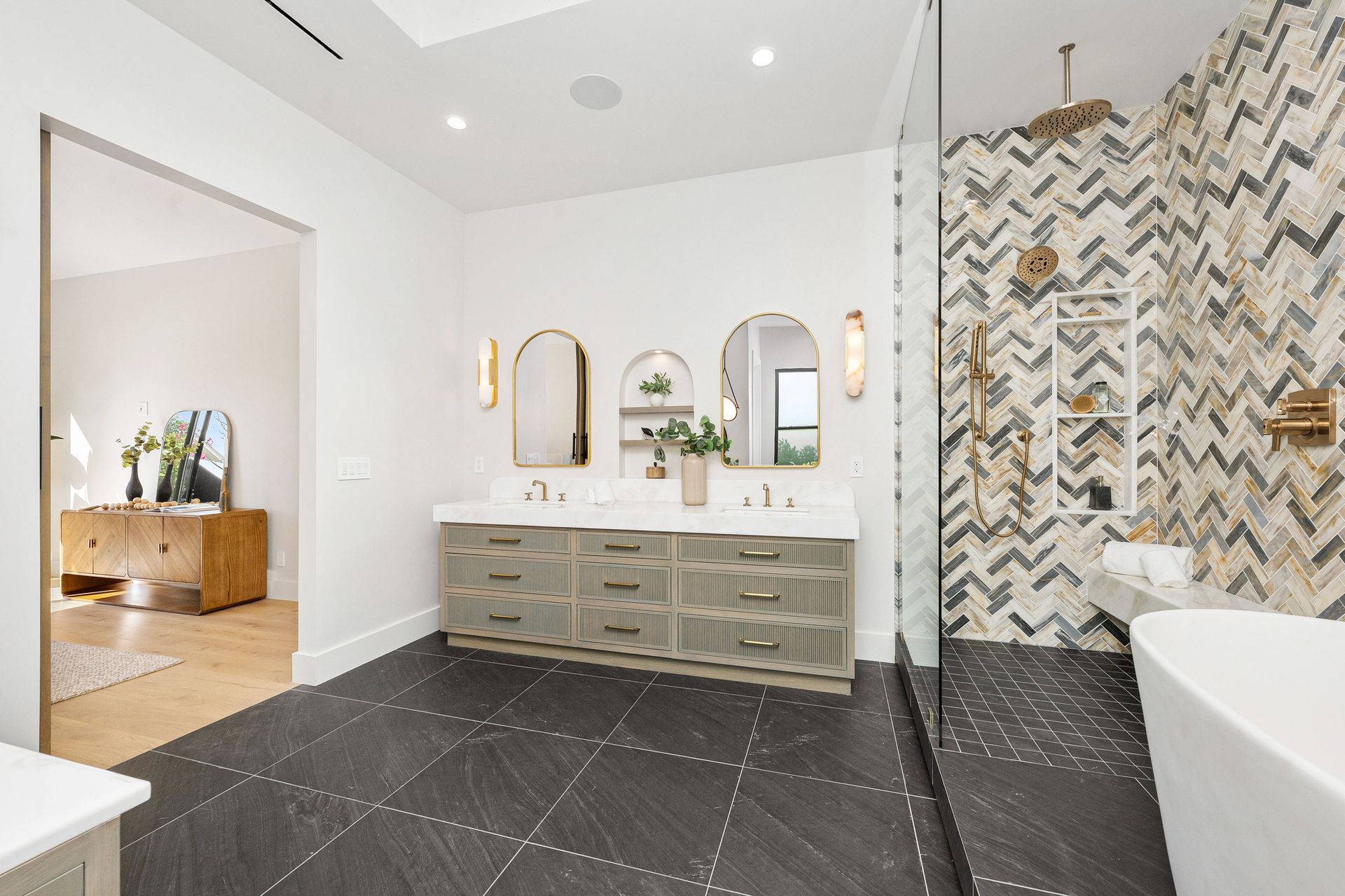 A bright, modern bathroom with a double vanity, arched mirrors, herringbone-tiled shower, and dark stone-patterned floor.