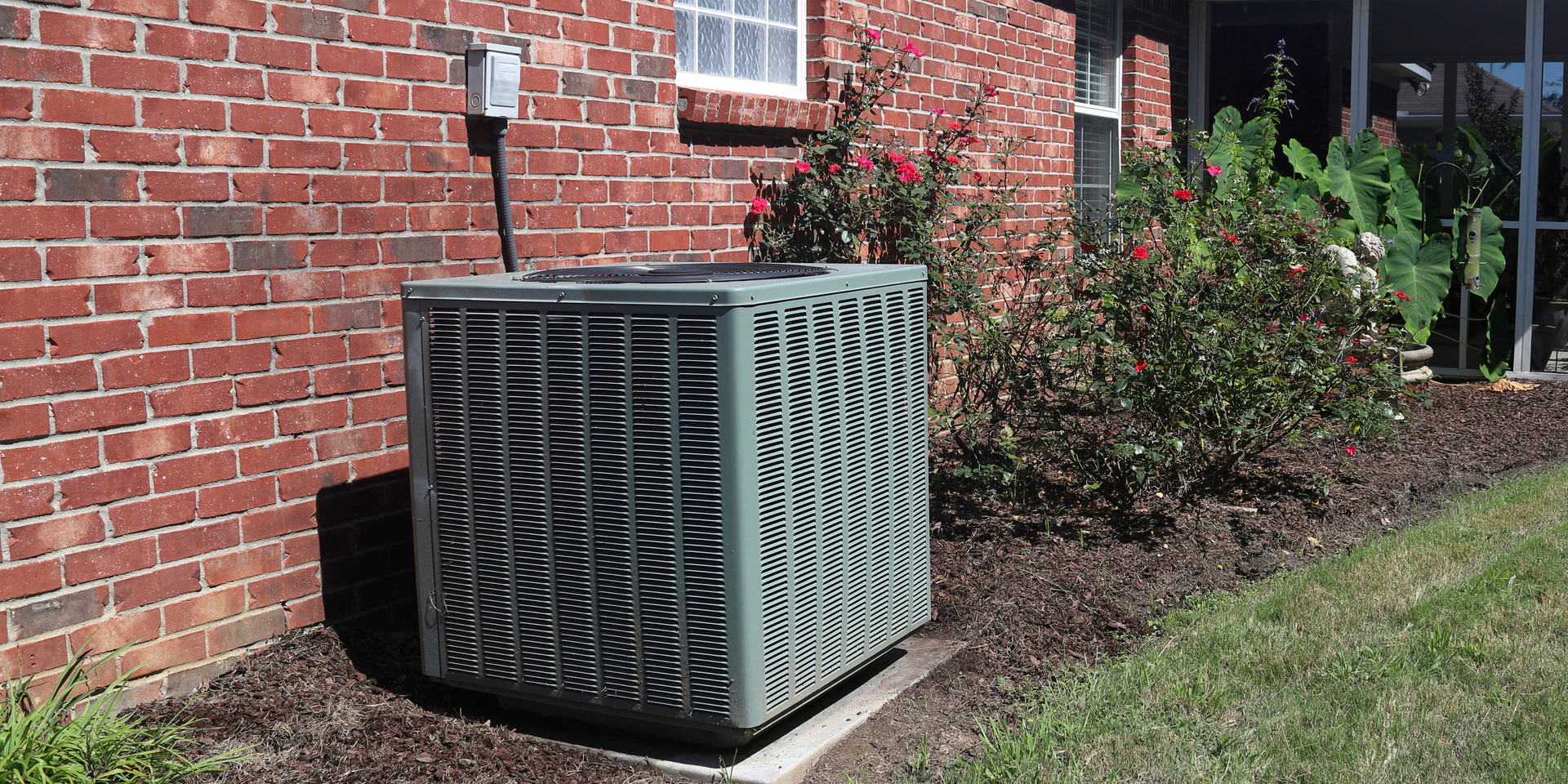 Outdoor HVAC unit beside a brick house, with a lawn and shrubs nearby